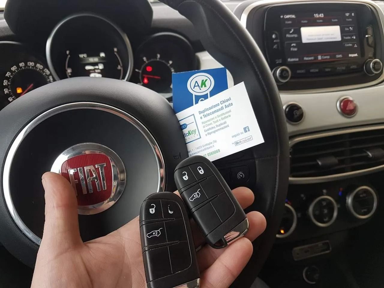 Mano che tiene due chiavi di un'auto davanti al volante di una Fiat. Cruscotto interno visibile con la tessera 