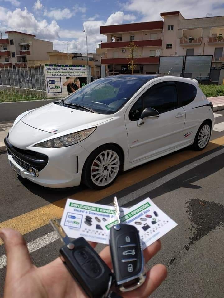 Una persona tiene in mano le chiavi di un'auto davanti a una Peugeot 207