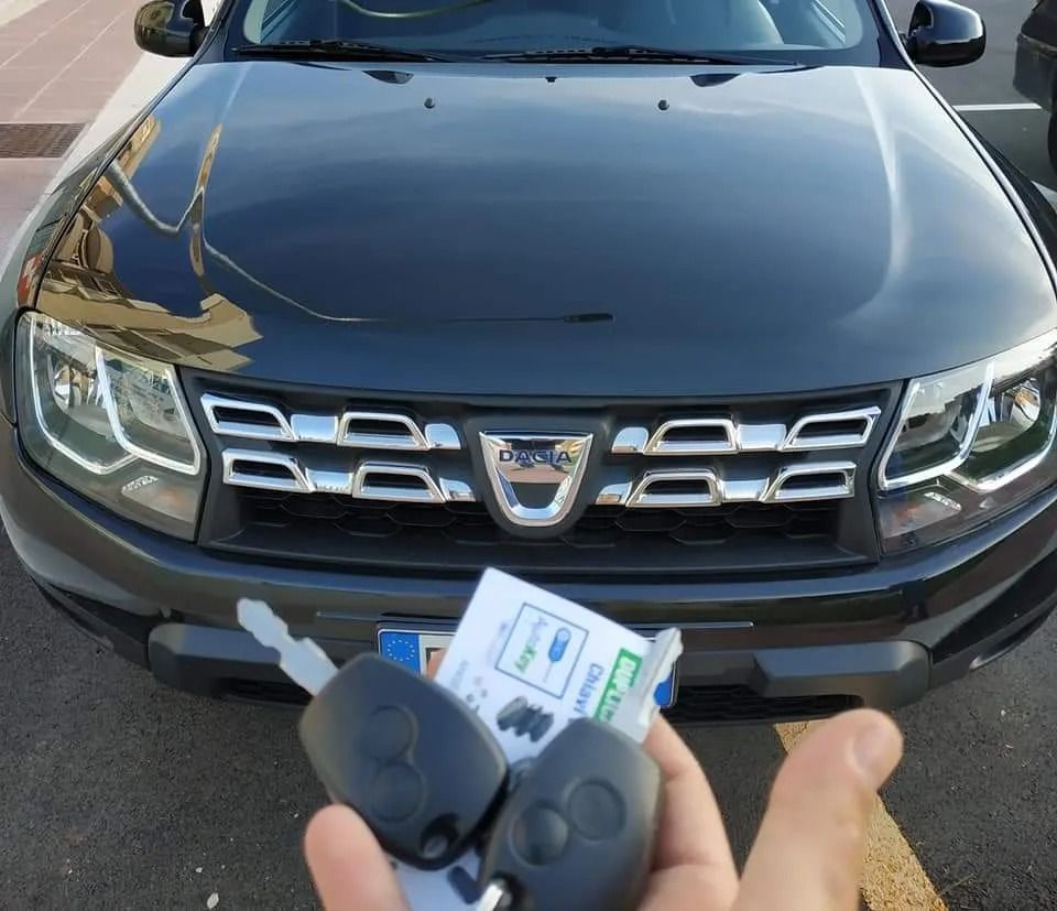Una persona tiene in mano due chiavi di un'auto davanti a una Dacia Duster grigio scuro, con un parcheggio sullo sfondo.