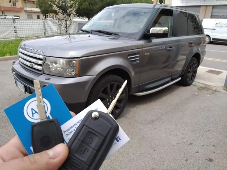 Una mano tiene le chiavi di un'auto davanti a una Range Rover grigia parcheggiata all'aperto. 