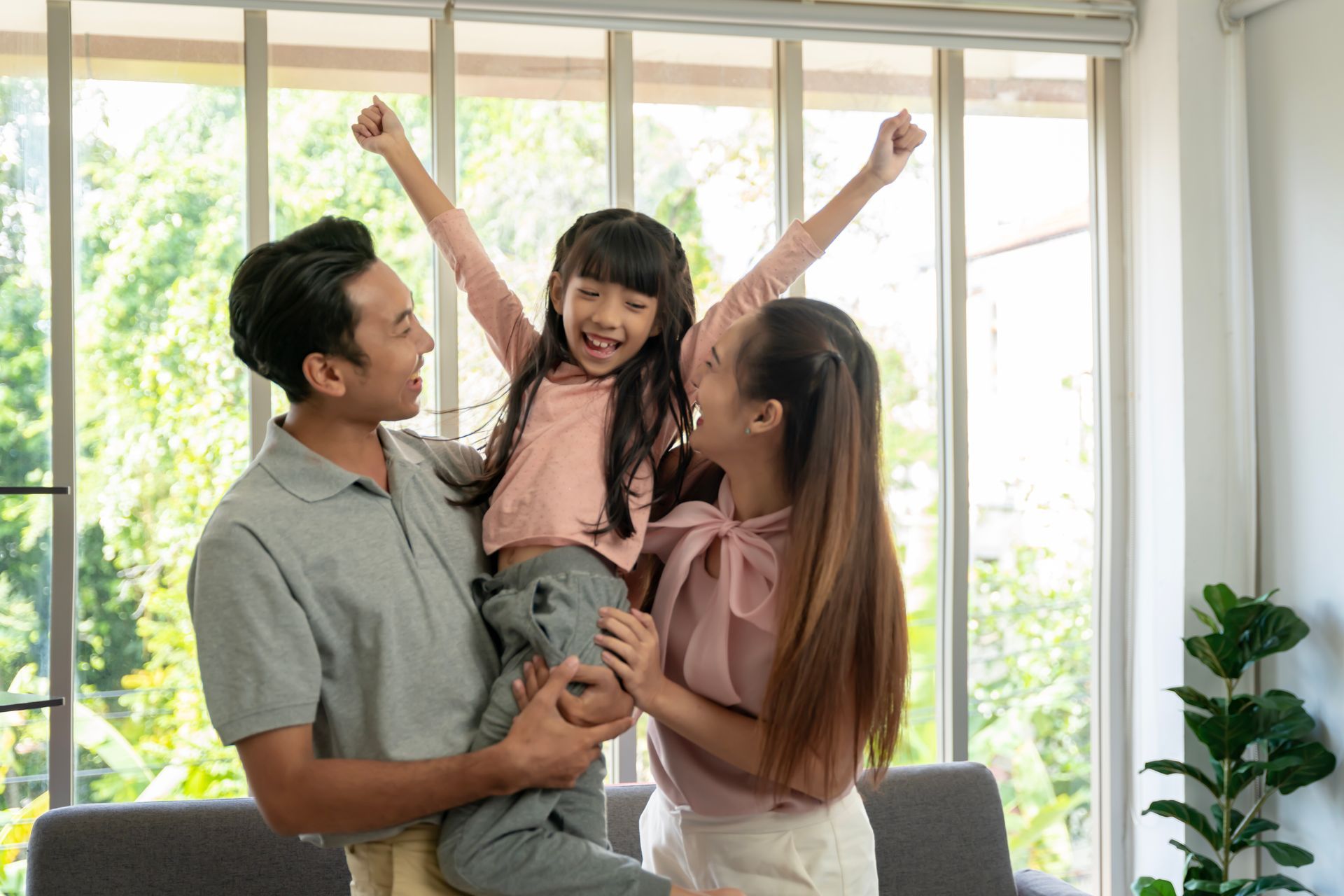 Happy family celebrating, holding daughter up. Smiling, raising arms in room with large window.