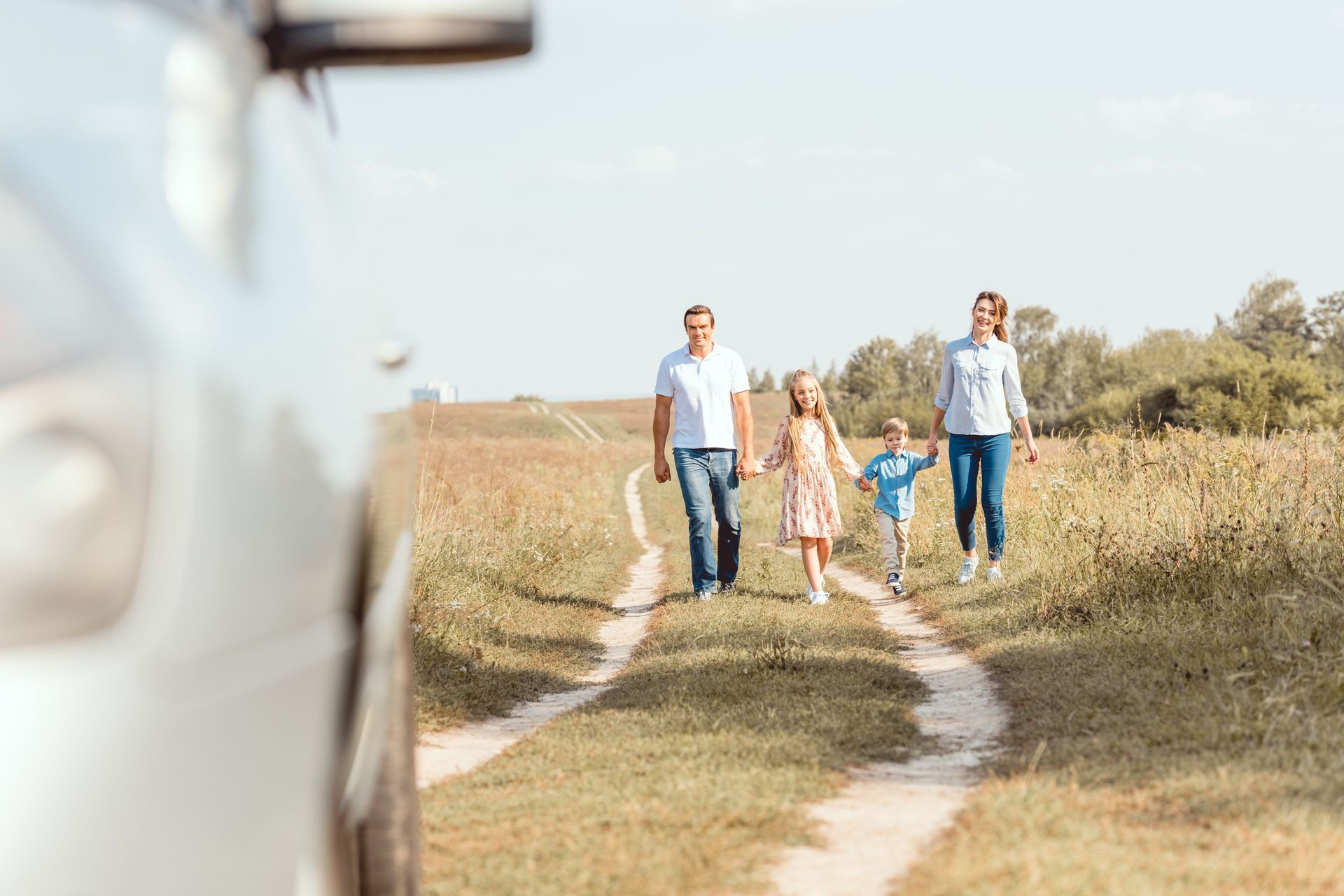 Family of four walking on a dirt path in a grassy field on a sunny day. A car is in the foreground.