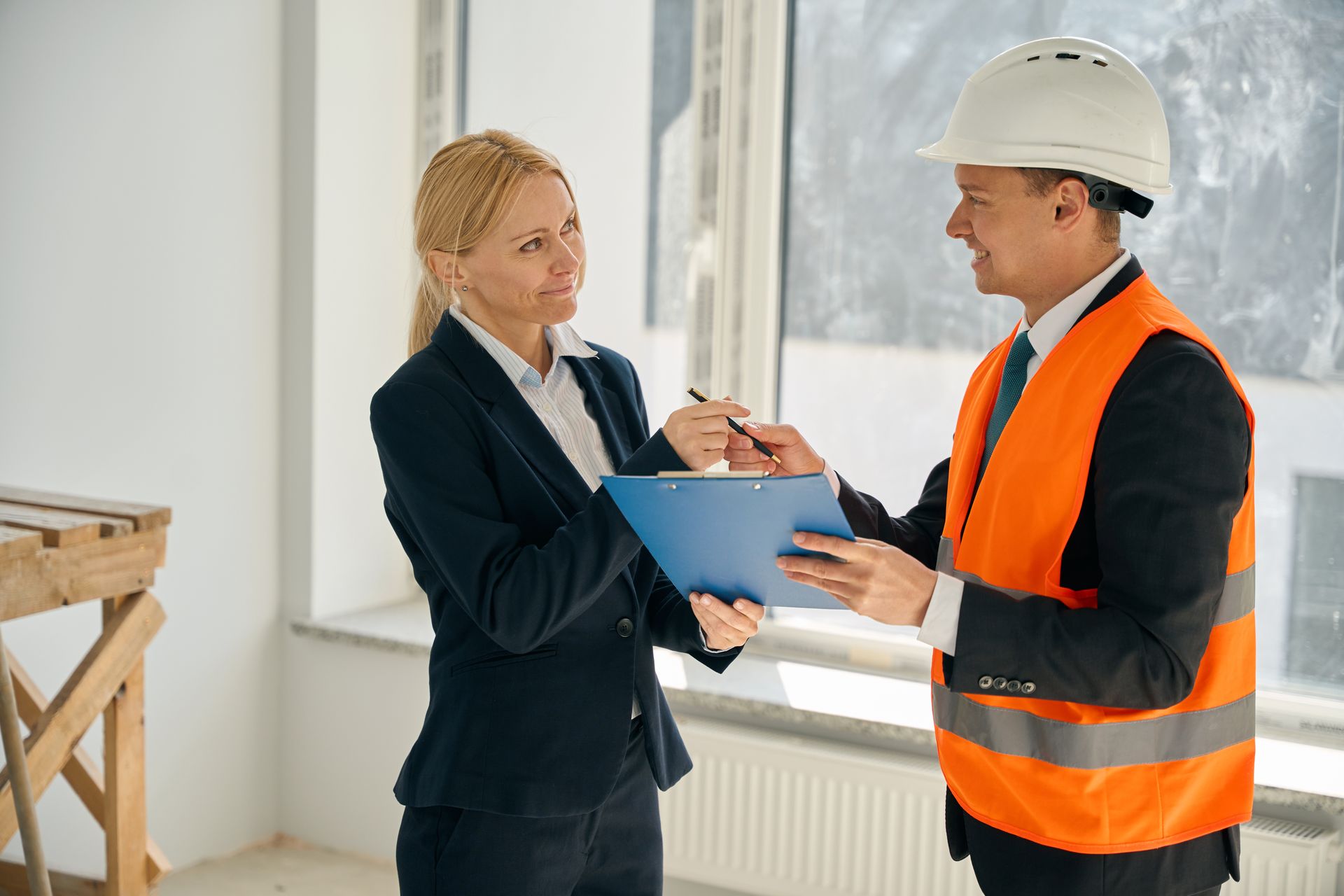 Woman in suit reviews clipboard with man wearing hard hat and safety vest by a window.