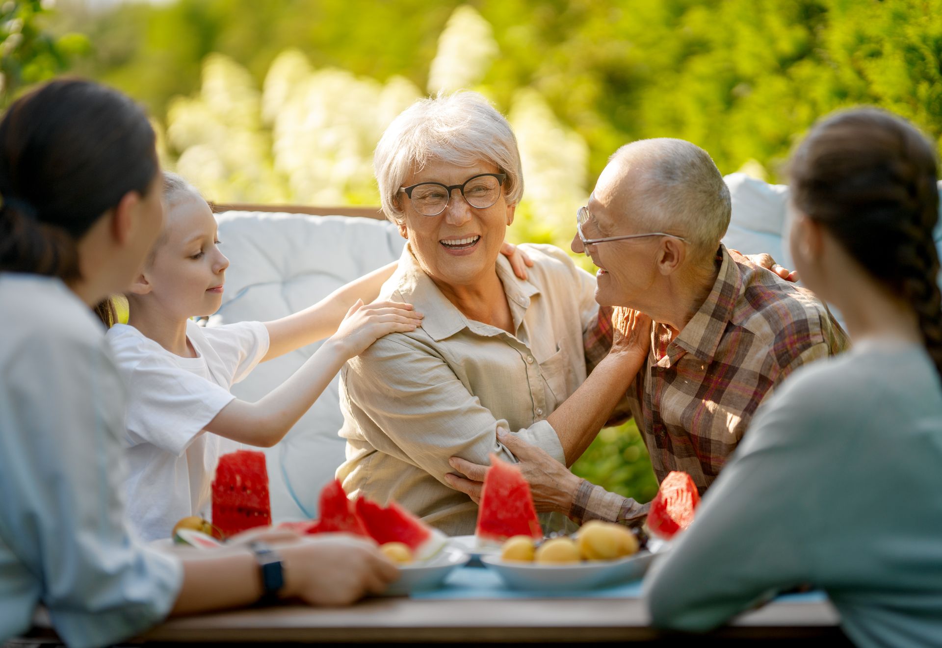 Family enjoying watermelon outdoors, smiling. Includes adults and a child at a table.