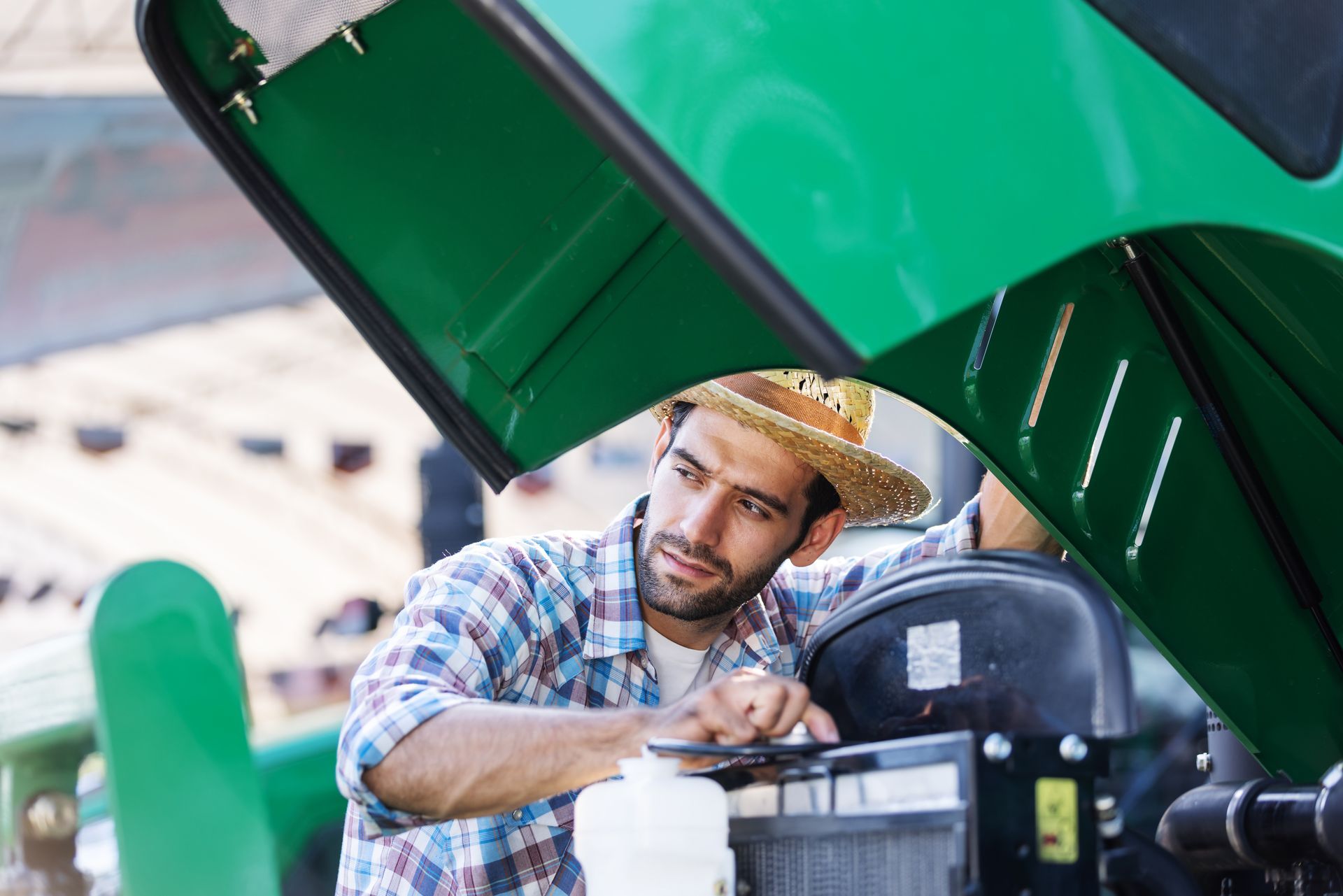 Man in straw hat checking tractor engine. Green tractor, open hood.