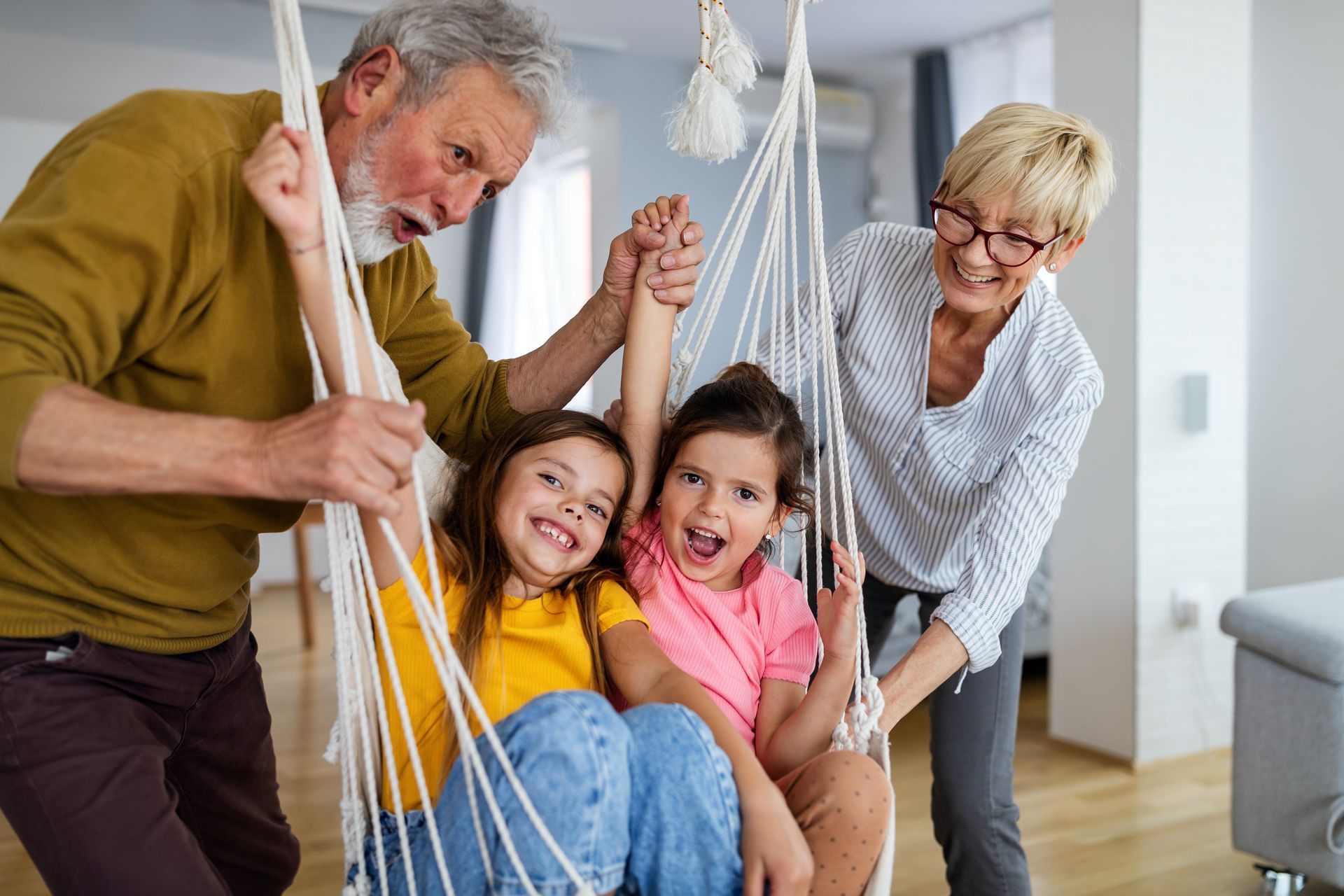Grandparents swing two young girls in an indoor chair; all are smiling and laughing.