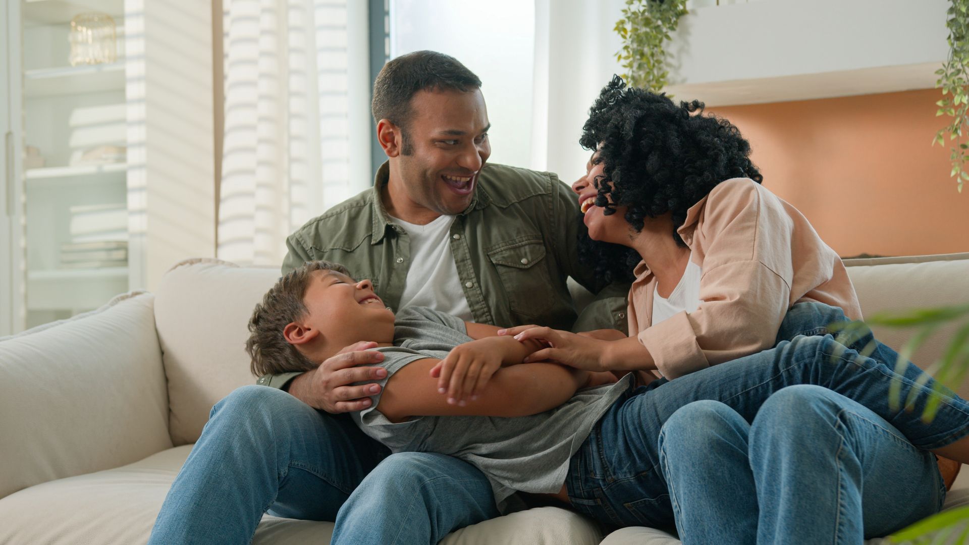 Family laughing together on a couch; mom and dad tickling child. Bright room, natural light.