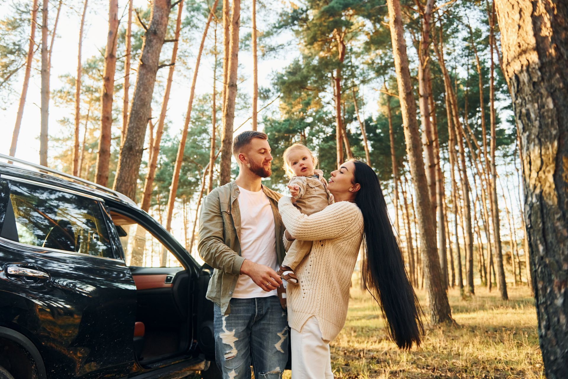 Family with a baby near a black SUV in a sunny forest setting.