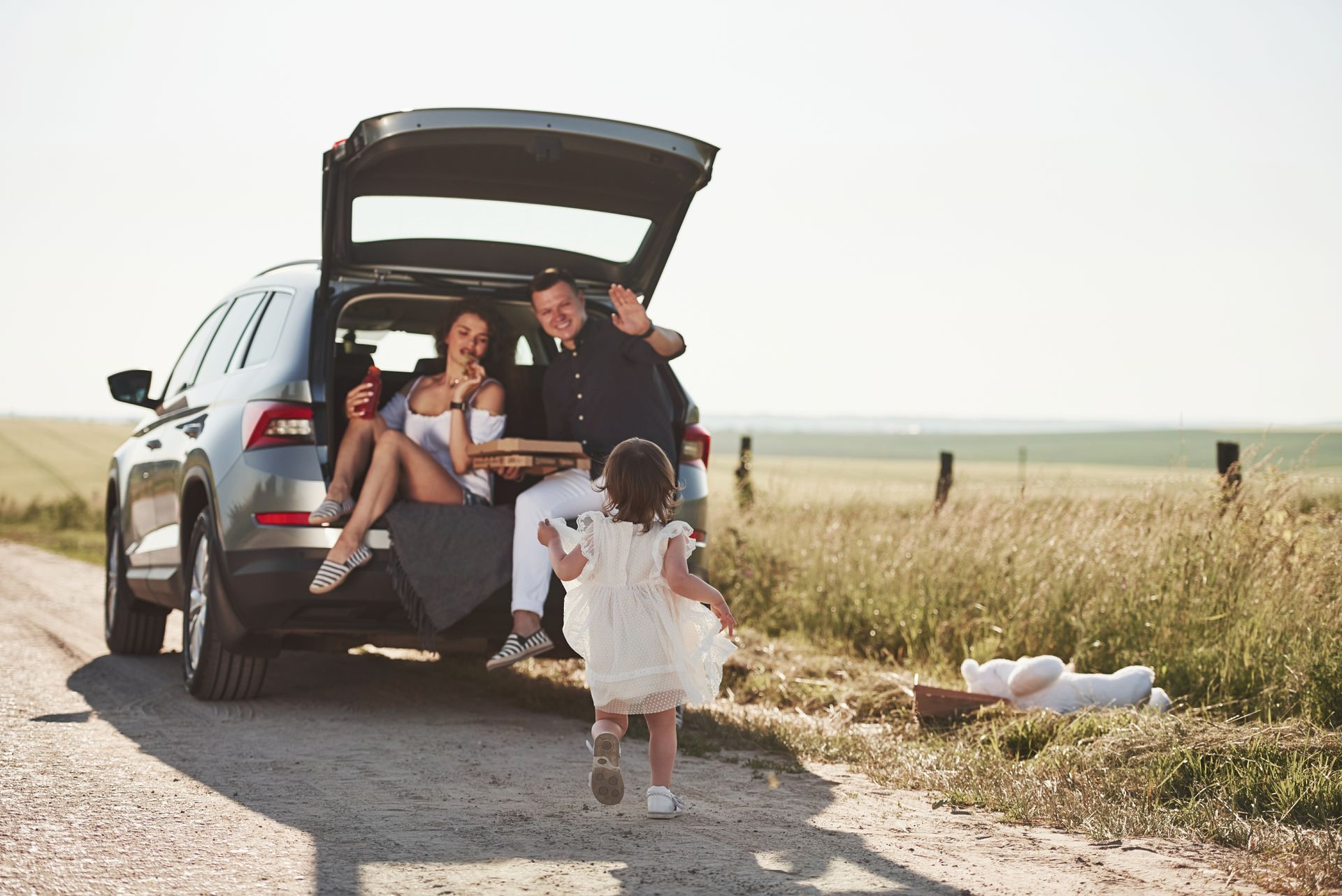 Family of four by an SUV with open trunk on a dirt road, child running toward them, sunny day.