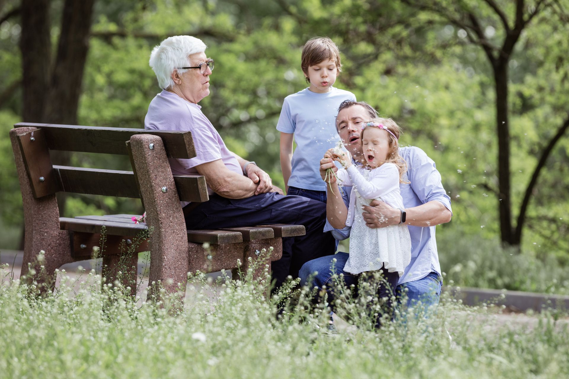 Family gathered at a park; an older person on a bench, a man holding a child with a dandelion, a boy standing behind.
