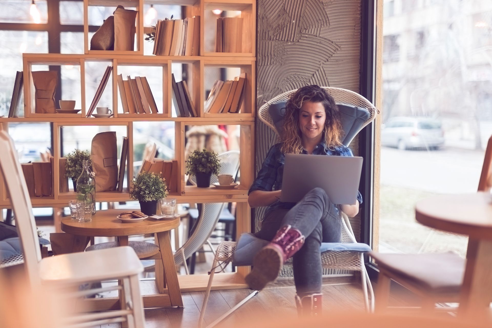 Woman sitting in a cafe, using a laptop, near a window and bookcase.