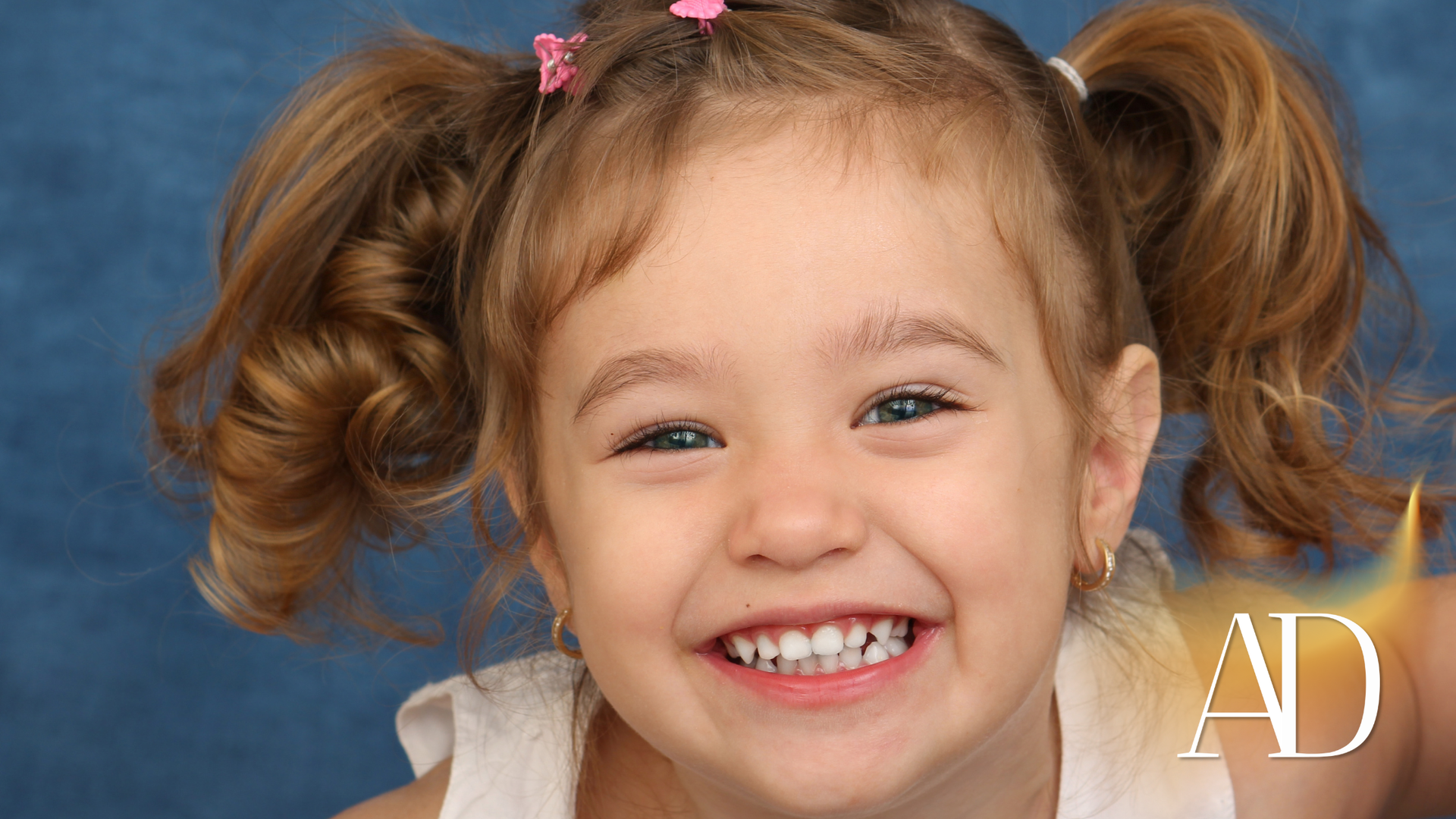 Smiling child with blonde pigtails, wearing a white top, against a blue background.