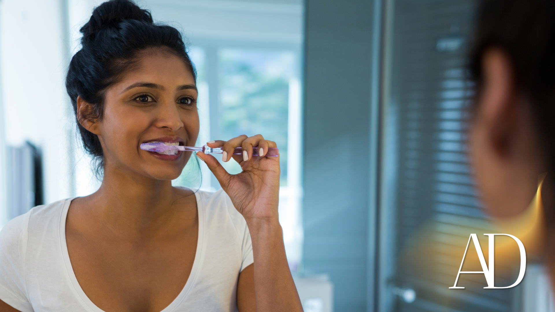 Woman brushing her teeth in front of a mirror, white top, light background.