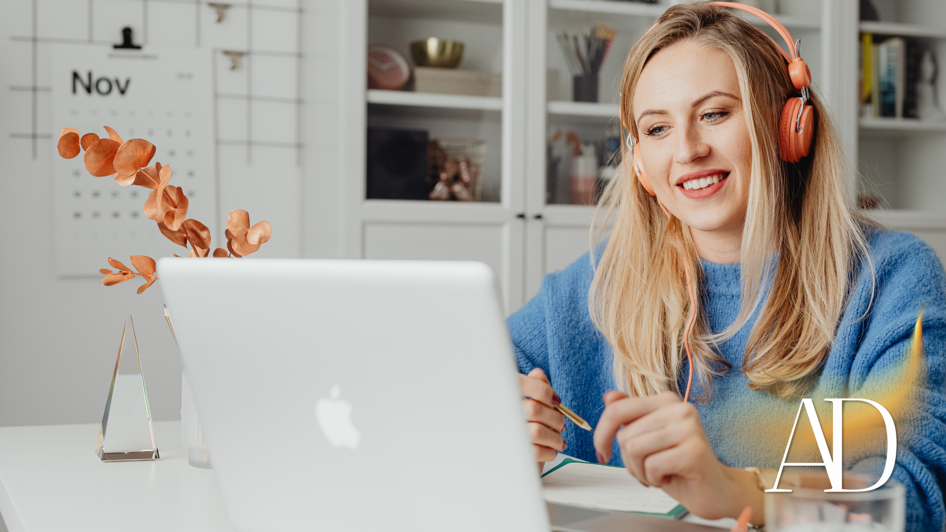 Woman wearing headphones smiles at a laptop, working at a white desk in a room.