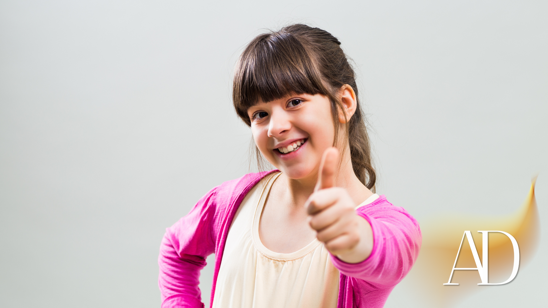 Girl in pink cardigan gives a thumbs up, smiling, against a gray background.