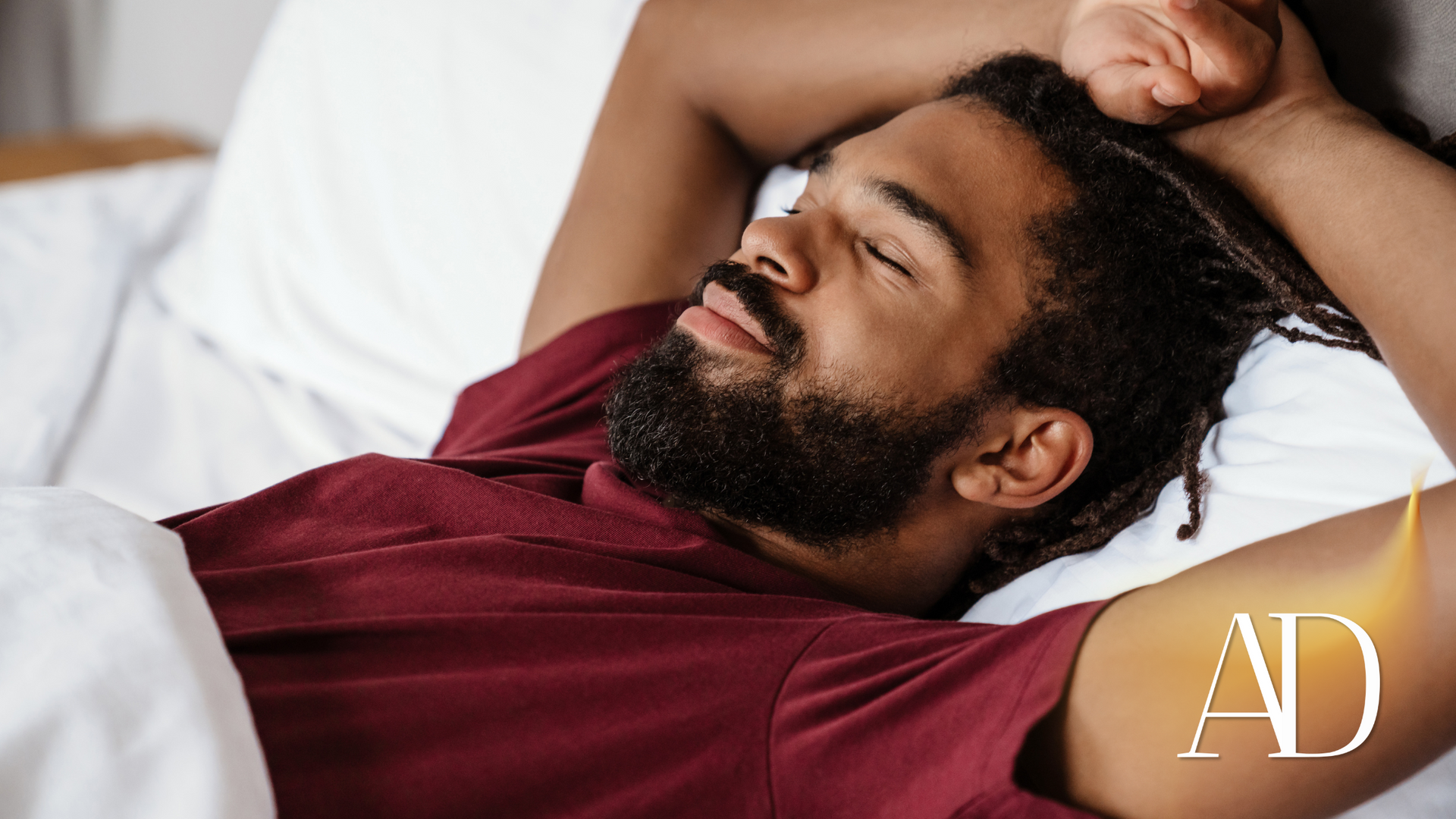 A man with a beard is sleeping in a bed with his arms outstretched.