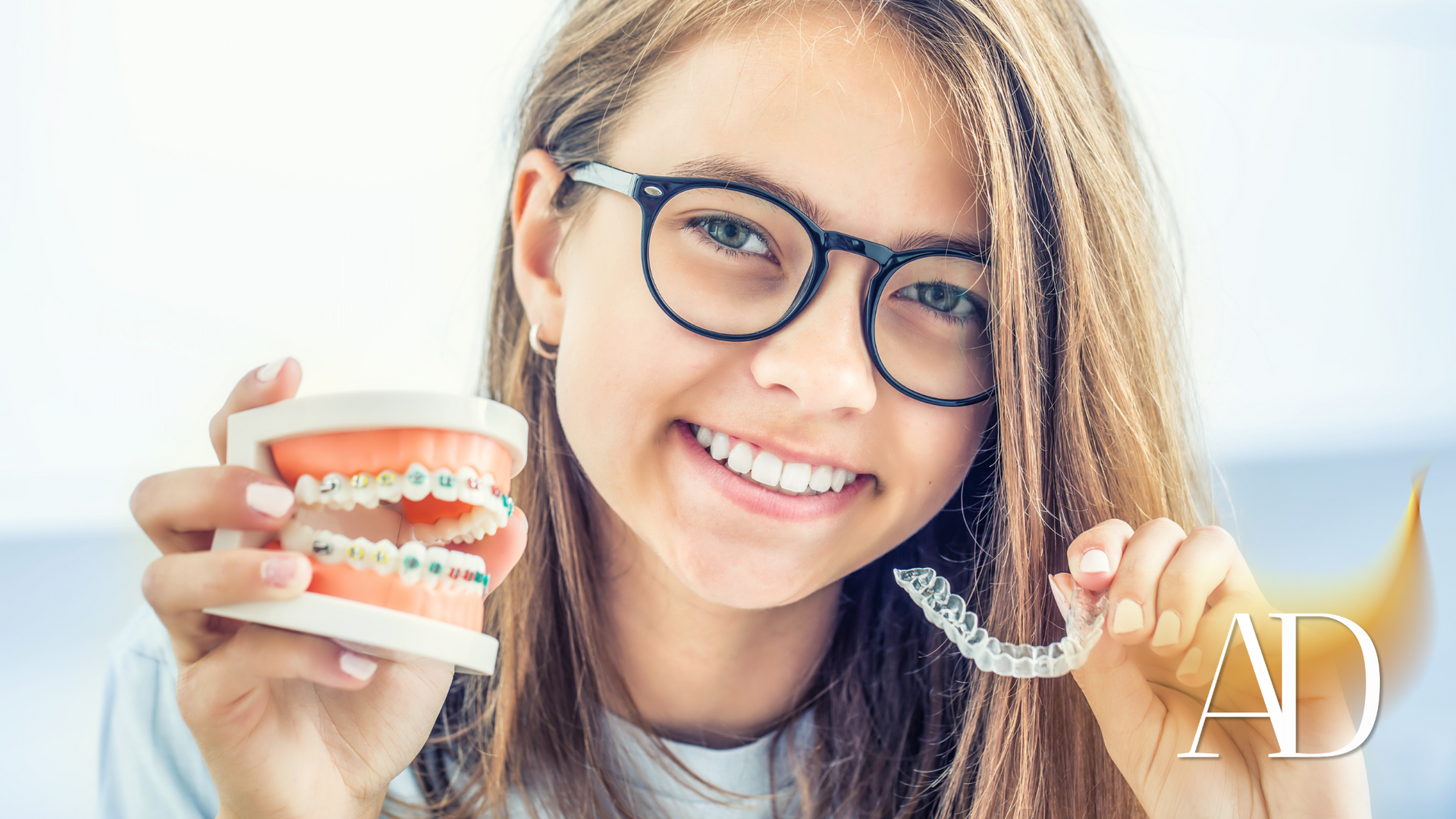 A young girl wearing glasses is holding a model of teeth and braces.