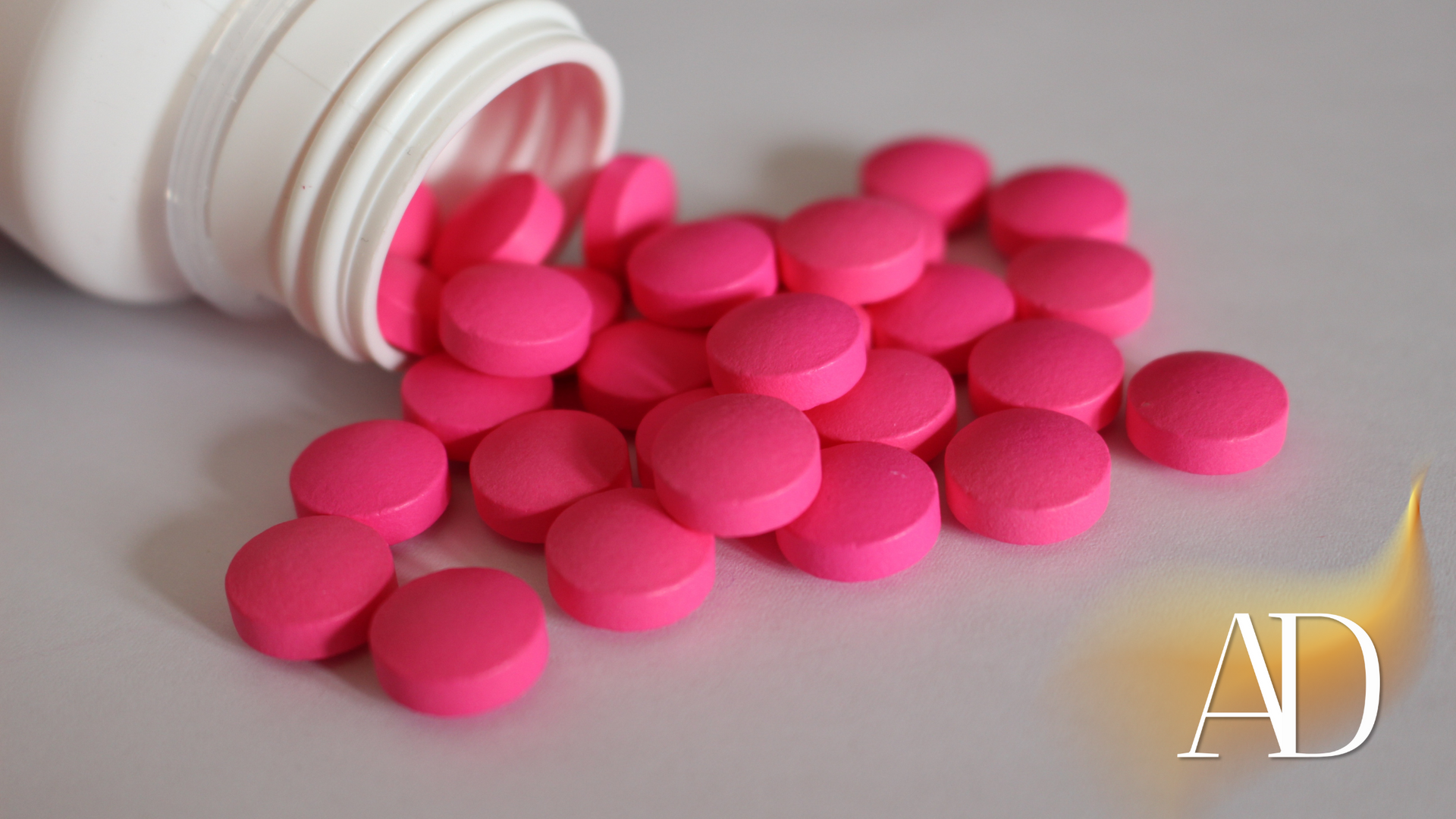 Pink pills are pouring out of a white bottle on a table.