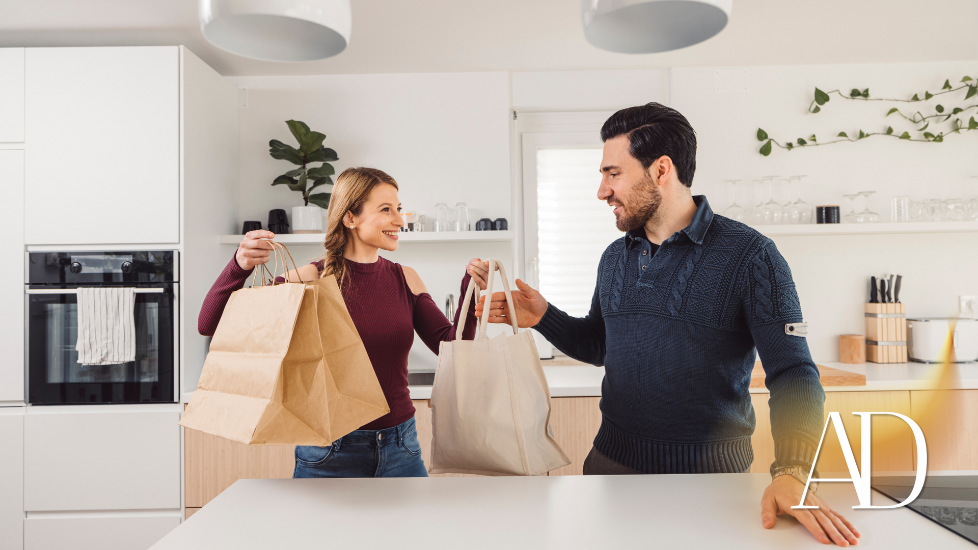 A man and a woman are standing in a kitchen holding shopping bags.