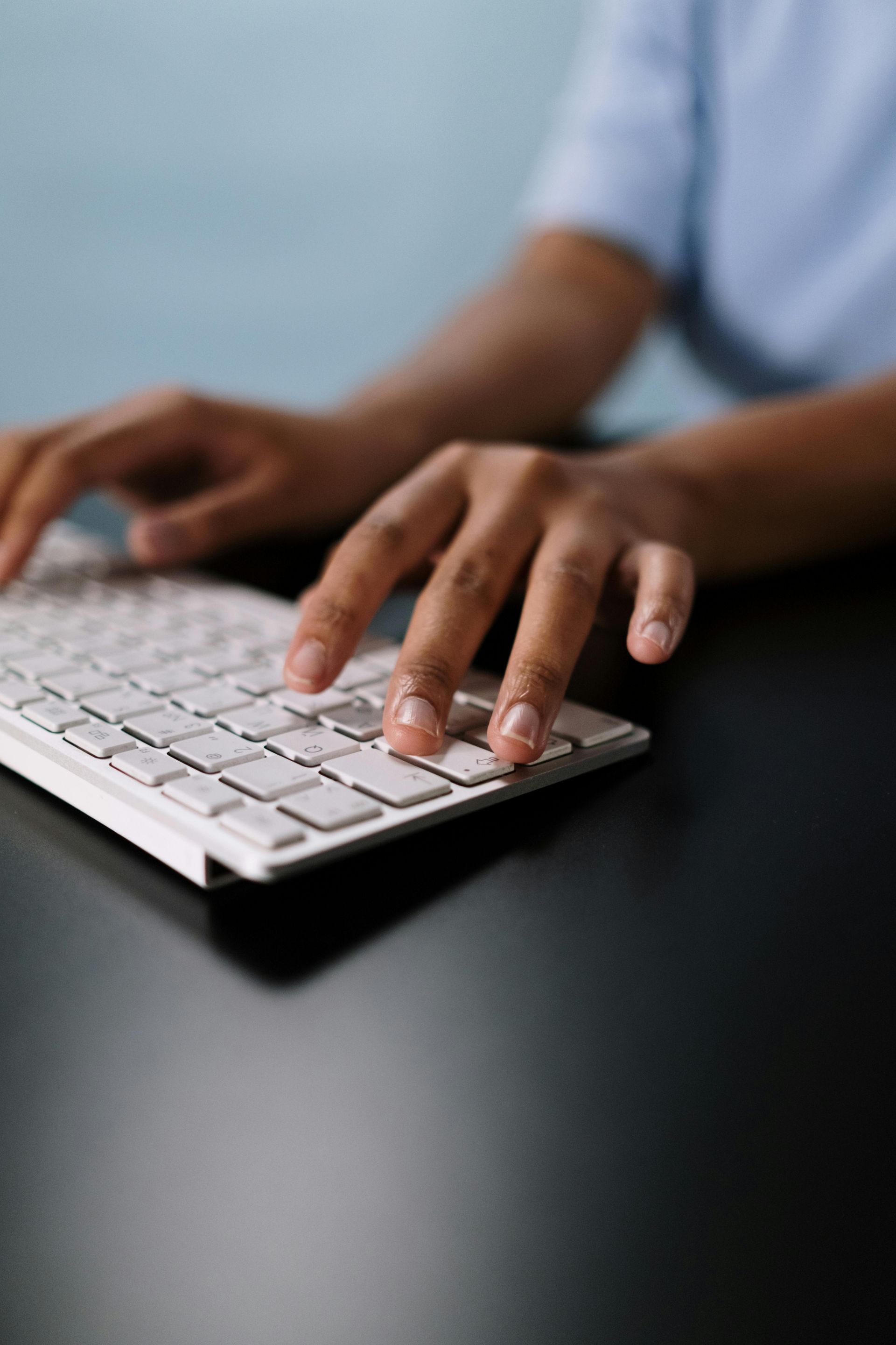 Hands typing on a white keyboard at a dark desk, with a blurred person in a blue shirt in the background