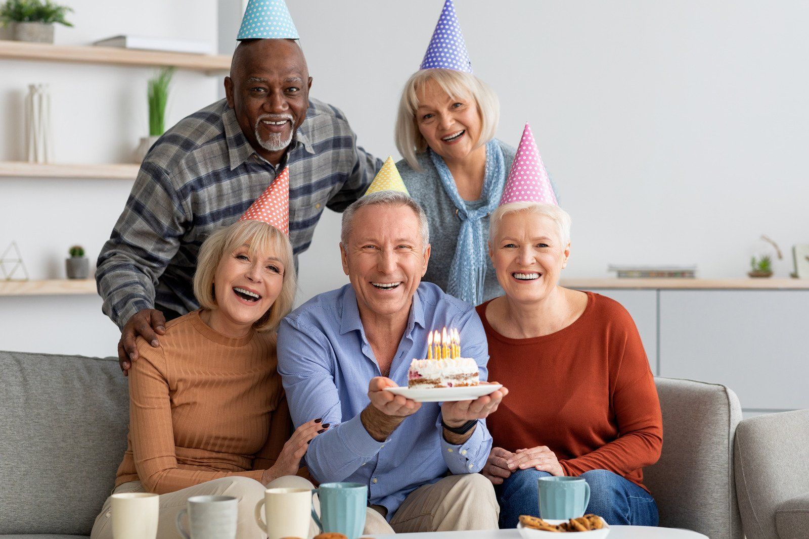 Group of people in party hats celebrating with a cake; smiles, indoors.