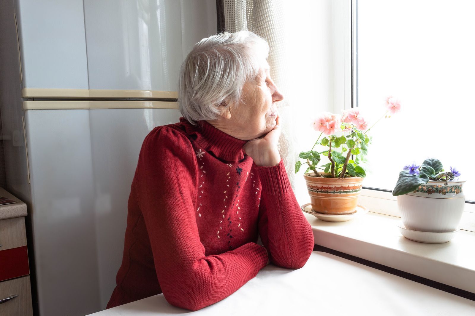 Woman in red sweater looking out a window, resting chin on hand. Flowers on windowsill.