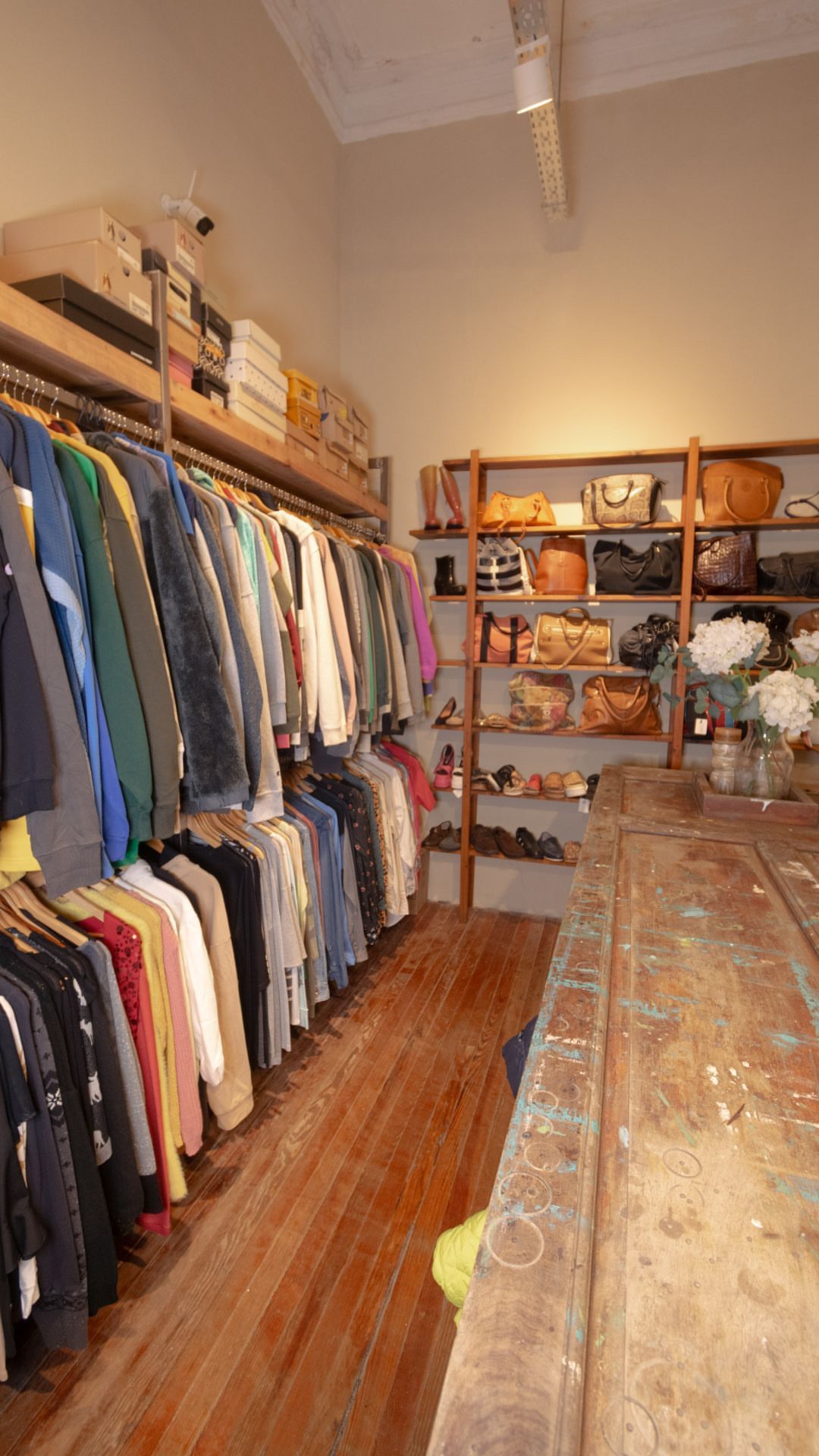 Pequeña boutique con percheros, estantes de madera y una larga mesa rústica en una habitación estrecha.