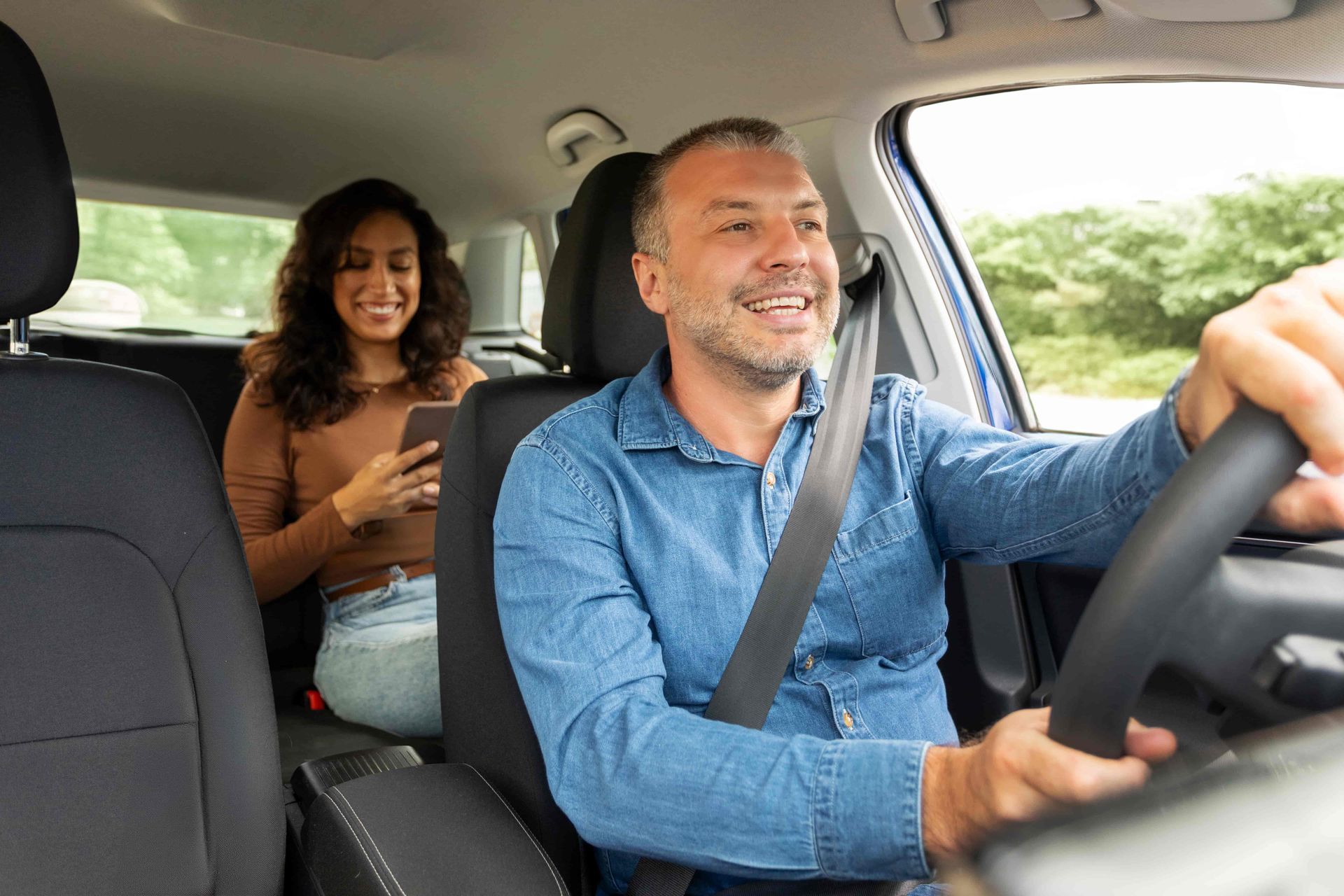 Man driving car, smiling, with passenger in backseat looking at phone. Interior car shot.