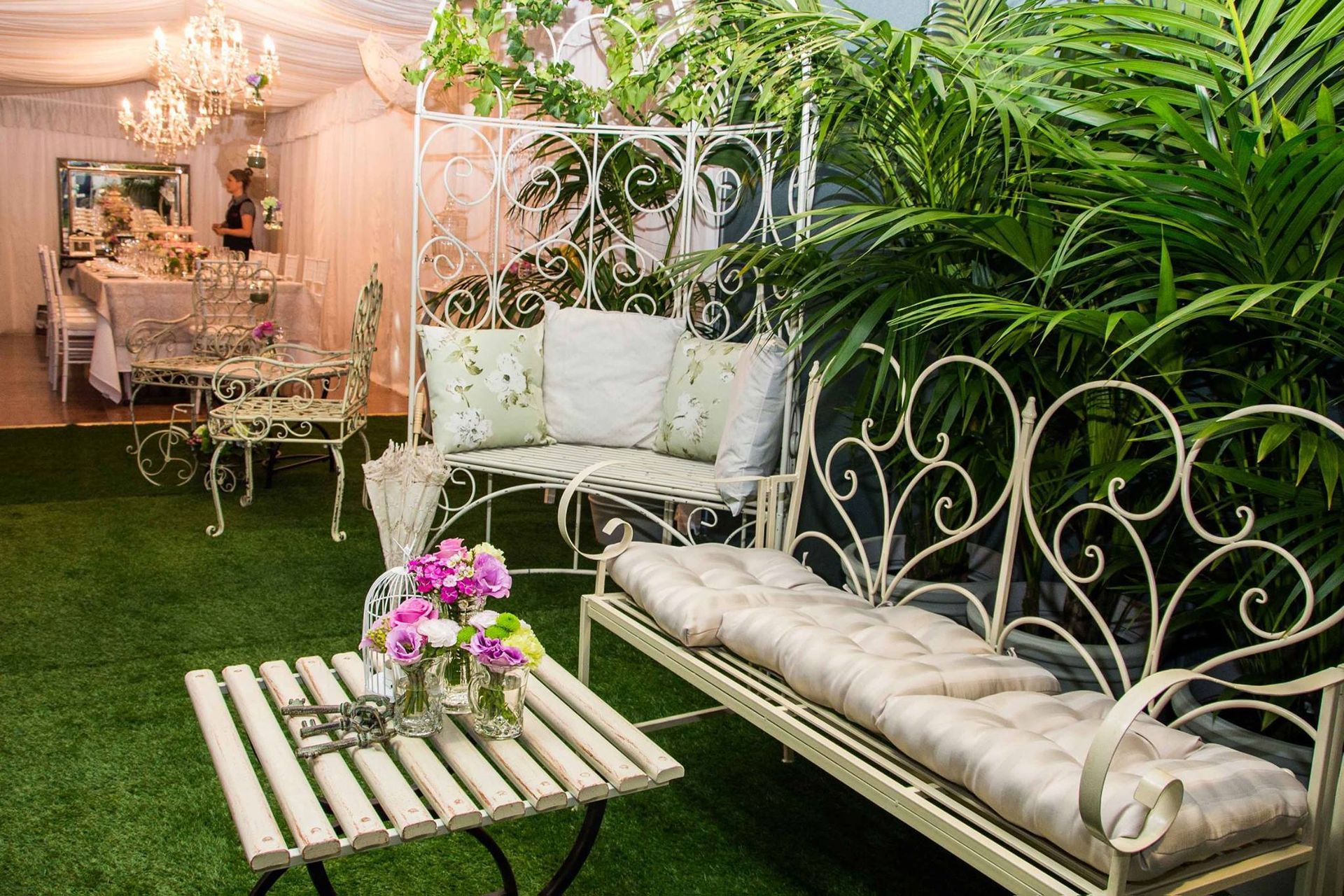A white wrought iron bench is sitting on top of a lush green lawn next to a table.
