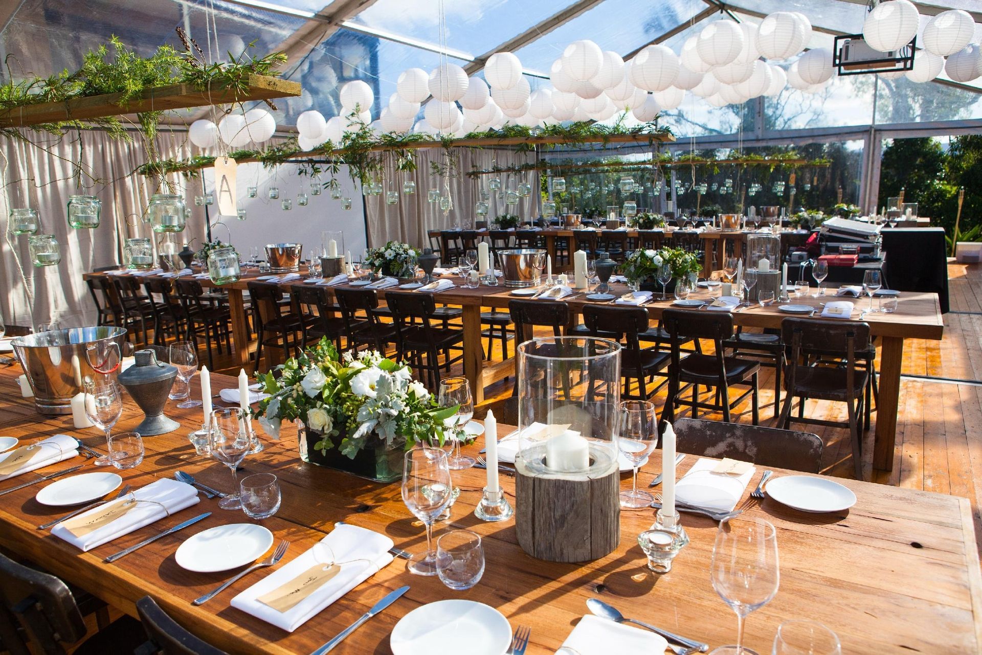 A long wooden table is set for a wedding reception under a clear tent.