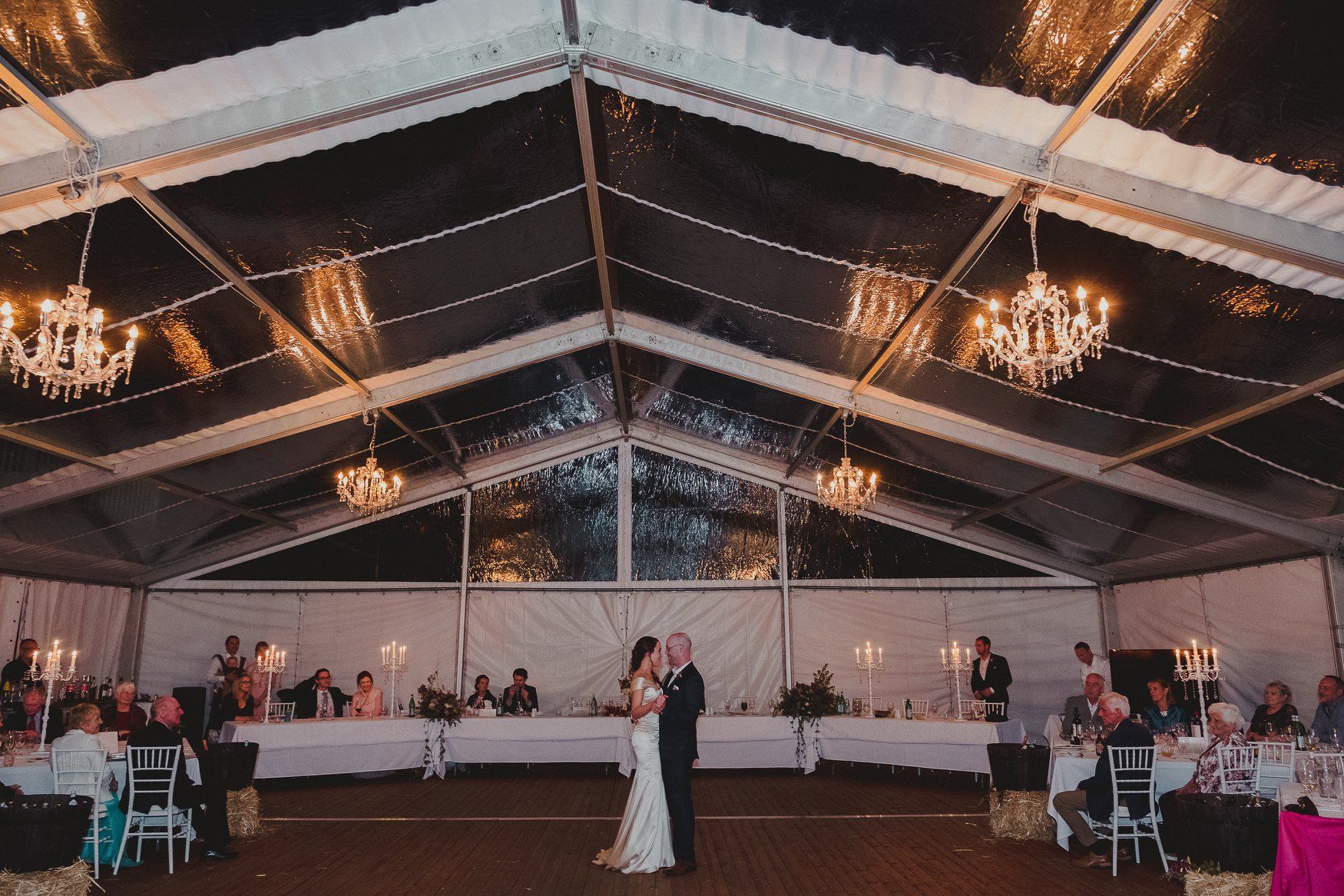 A bride and groom are dancing under a clear tent at their wedding reception.