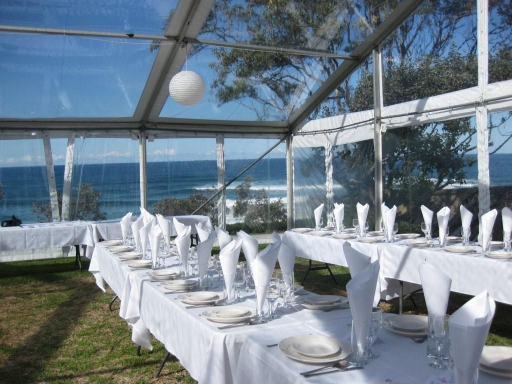 A long table is set under a clear tent with a view of the ocean.