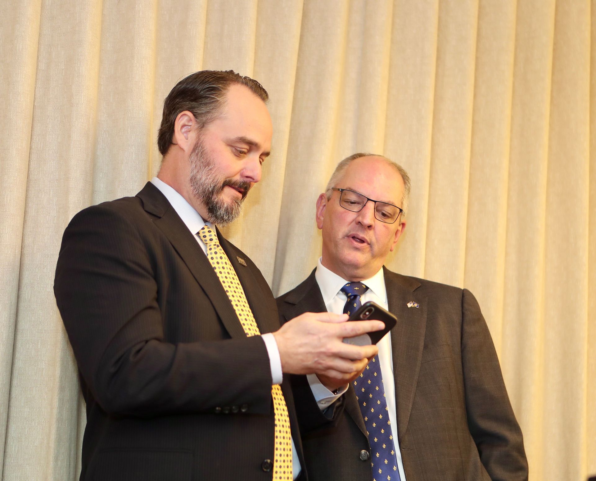 Two men in suits look at a smartphone, next to a beige curtain.
