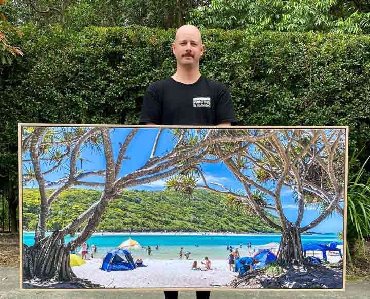 A Man is Holding a Large Painting of a Beach — Gold Coast Printing & Framing in Burleigh Heads, QLD
