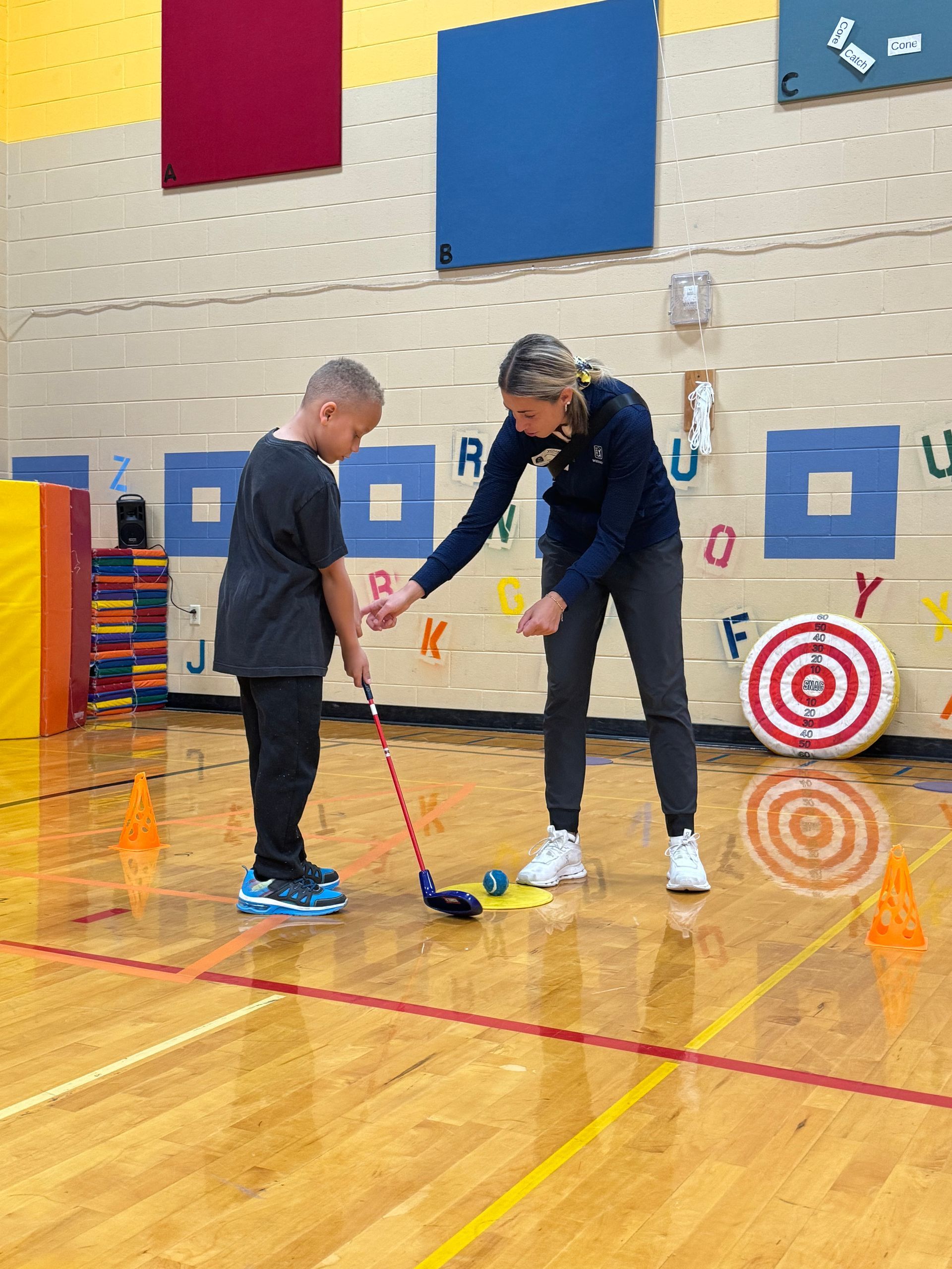 A woman is teaching a young boy how to play golf in a gym.