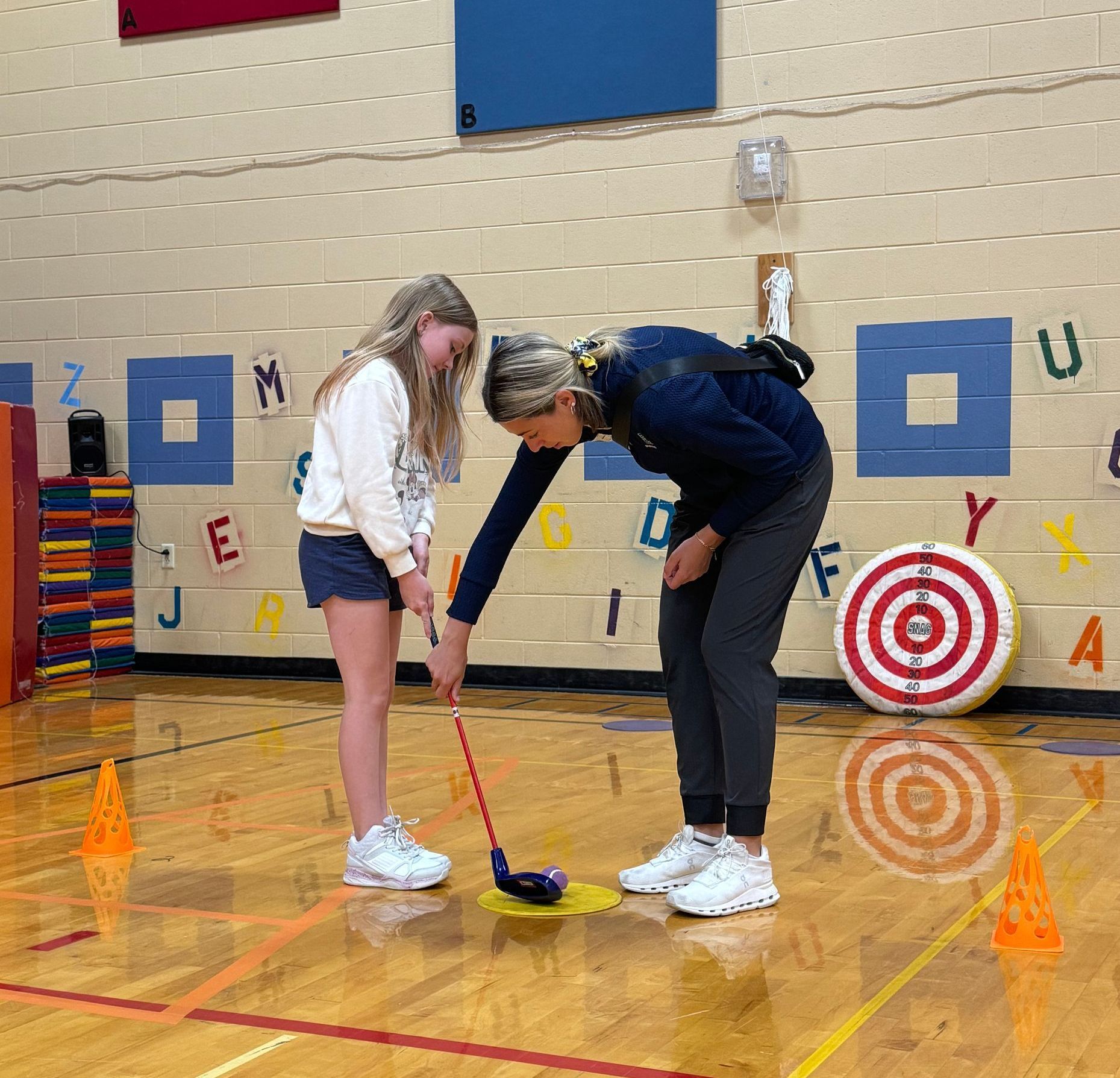 A woman is teaching a young girl how to play golf in a gym.