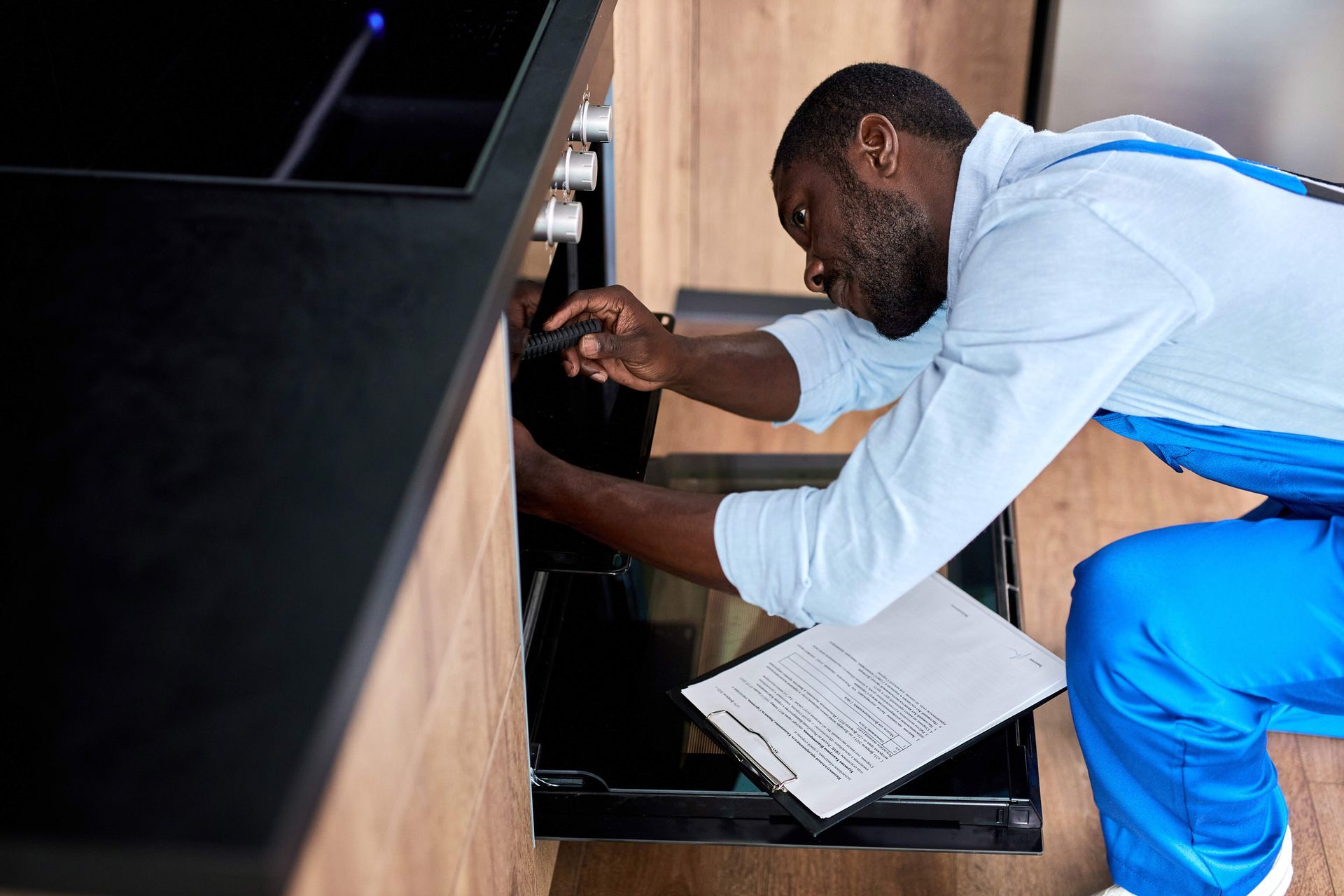 A person in a blue jumpsuit repairs an oven, holding a document, in a modern kitchen.