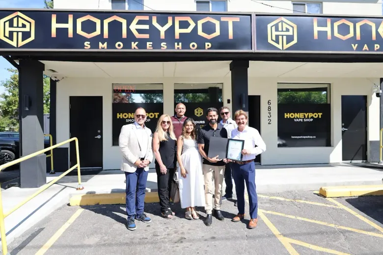A group of six people stand outside the Honeypot Smokeshop, holding a framed certificate in front of the storefront.