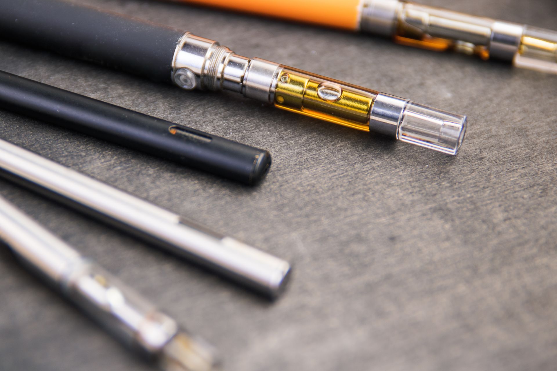 A close-up of several vape pens and cartridges with metallic, black, and orange finishes arranged on a gray surface.