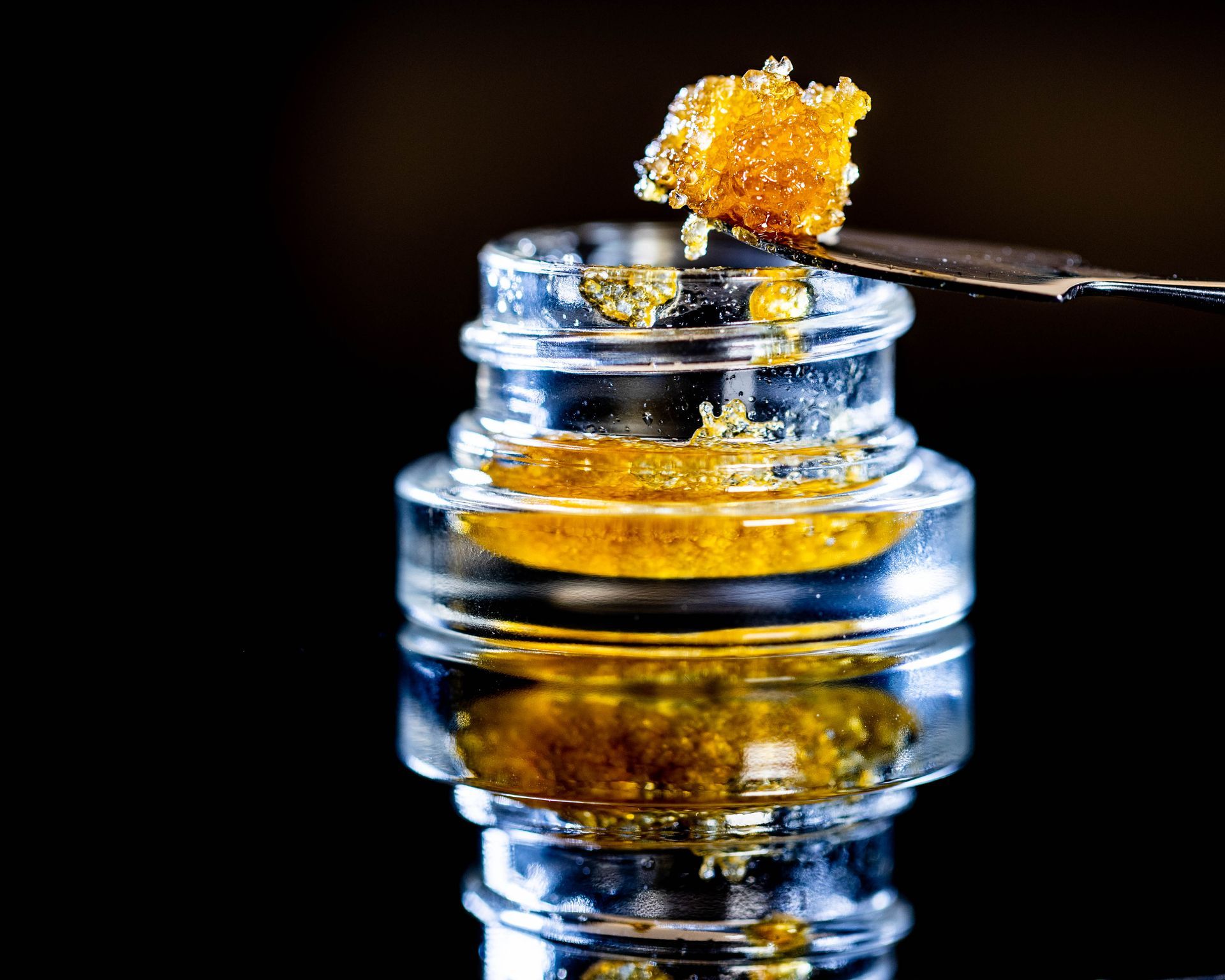 A metal dab tool holds a yellow cannabis concentrate over a small glass jar filled with the same golden substance.