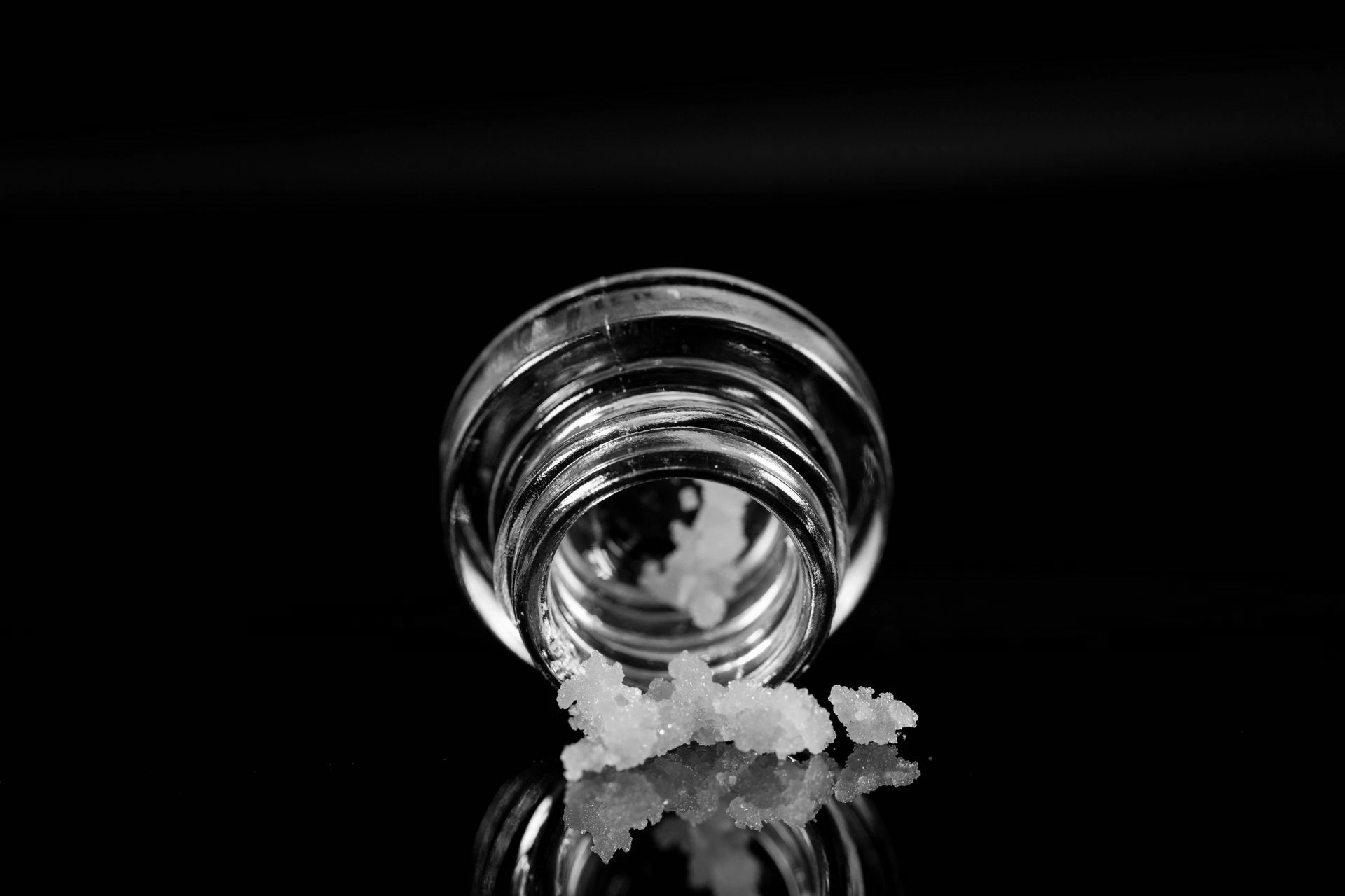 A small, open glass jar rests on a black surface, surrounded by white, crystalline granules.