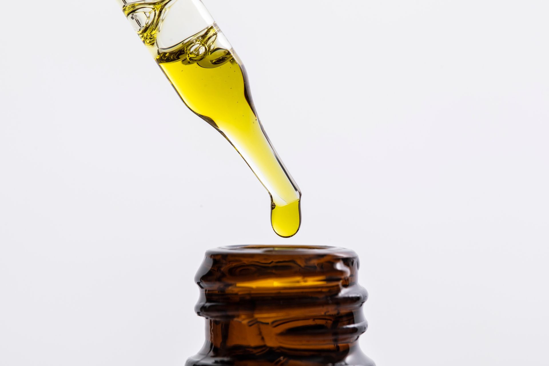 A glass pipette drips golden yellow liquid into the open mouth of a brown glass apothecary bottle against a white backdrop.