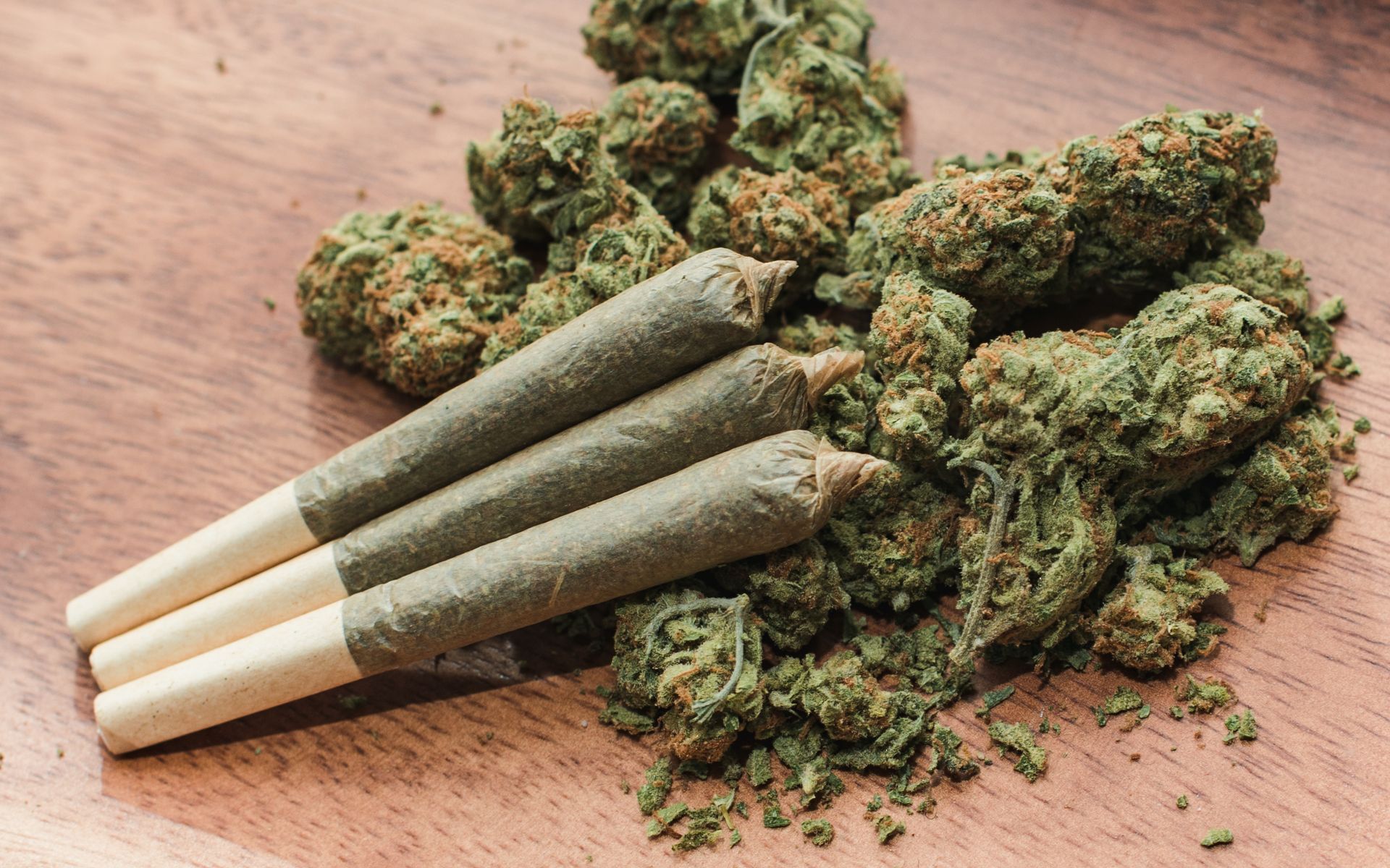 Three hand-rolled cannabis joints resting on a pile of dried cannabis flower buds against a wood-grain surface.