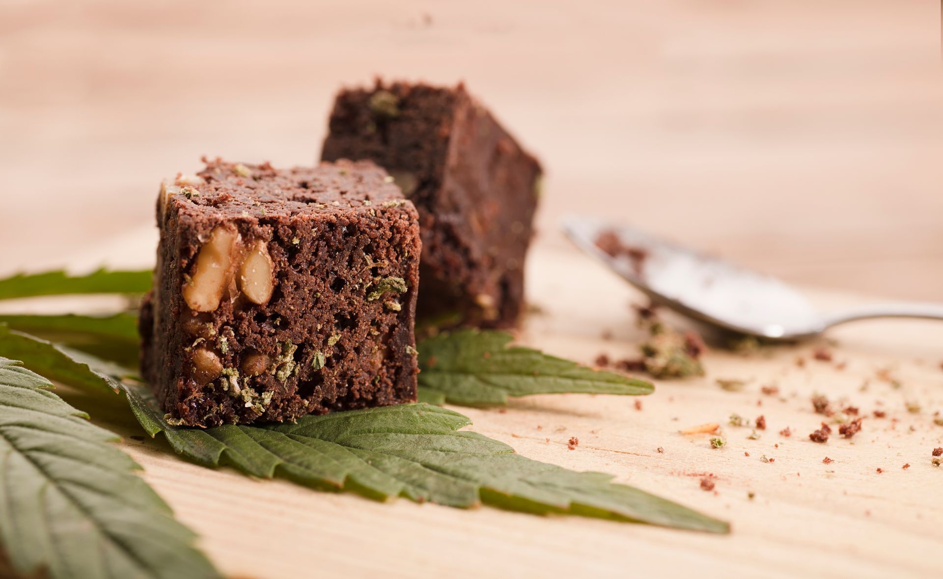 Two square chocolate walnut brownies garnished with marijuana leaves on a wooden cutting board with a spoon.