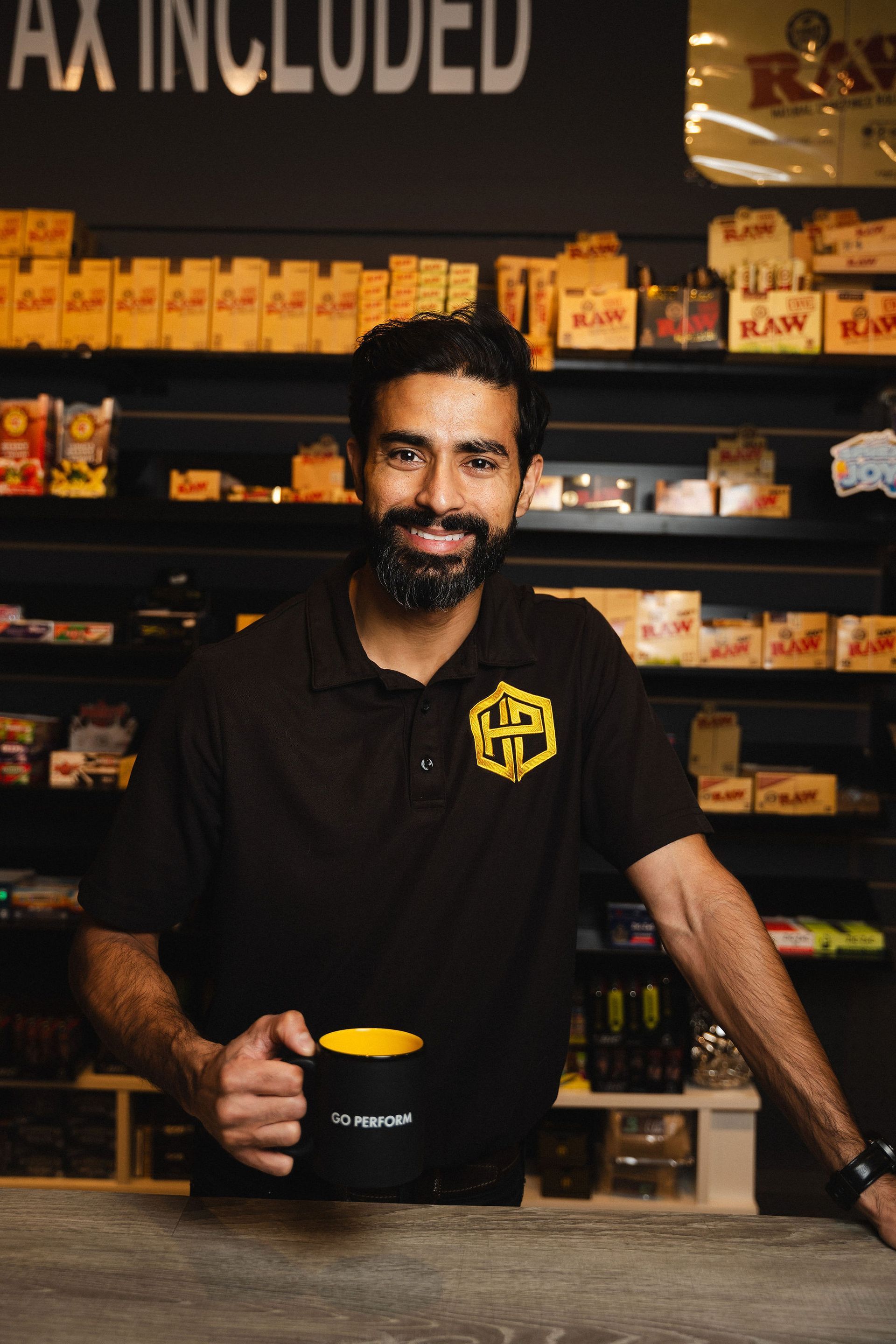 A person in a black polo shirt smiles while holding a black and yellow mug in a shop with shelves of merchandise.