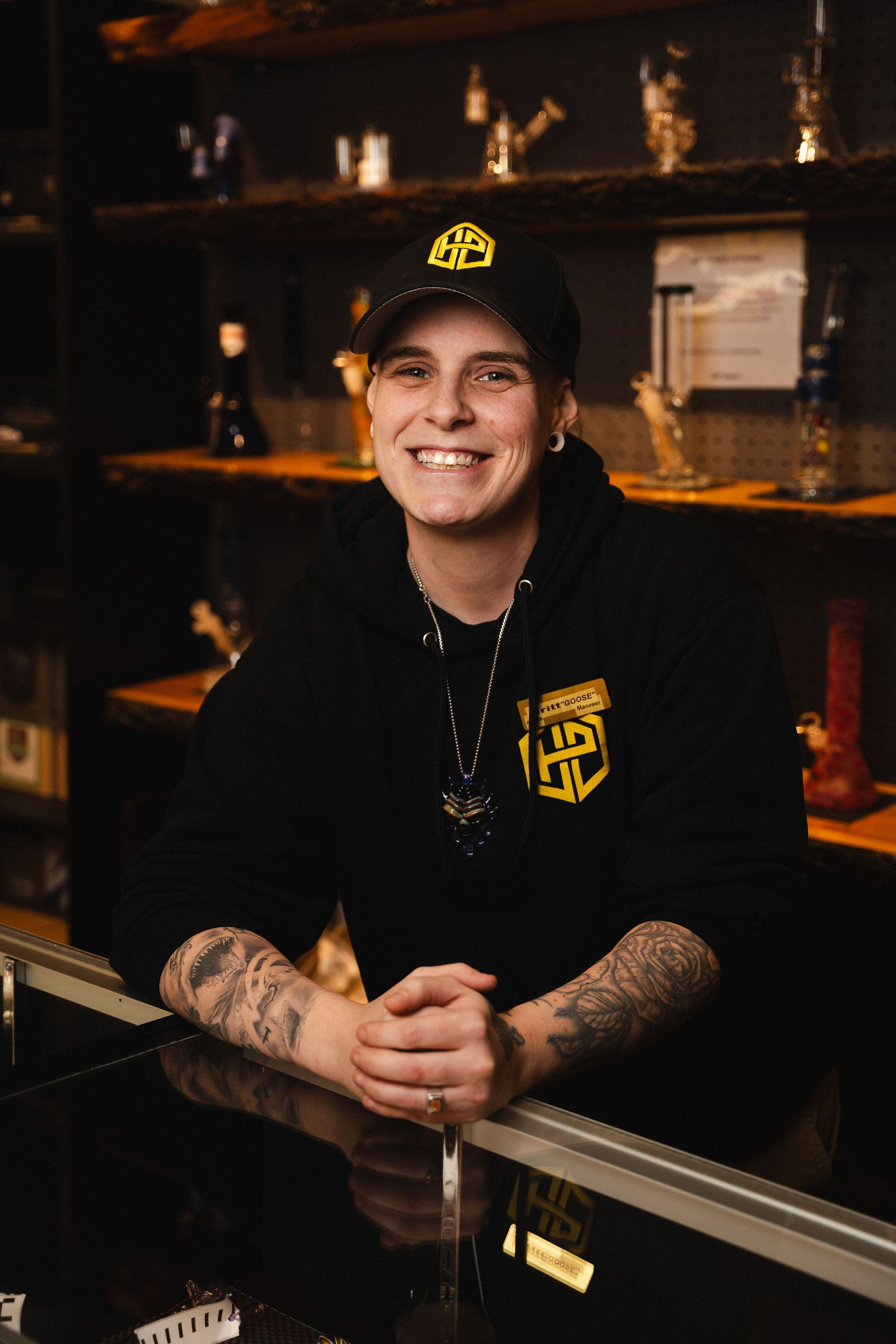 A smiling person in a black hoodie and cap stands behind a retail counter at a niagara falls dispensary, with shelves of glass products behind them.