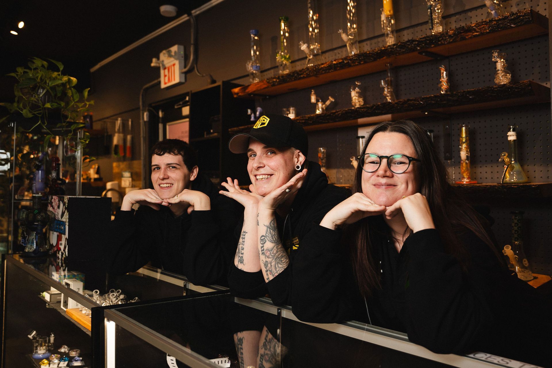Three people wearing black sweatshirts smile with their chins resting on their hands behind a glass store counter.