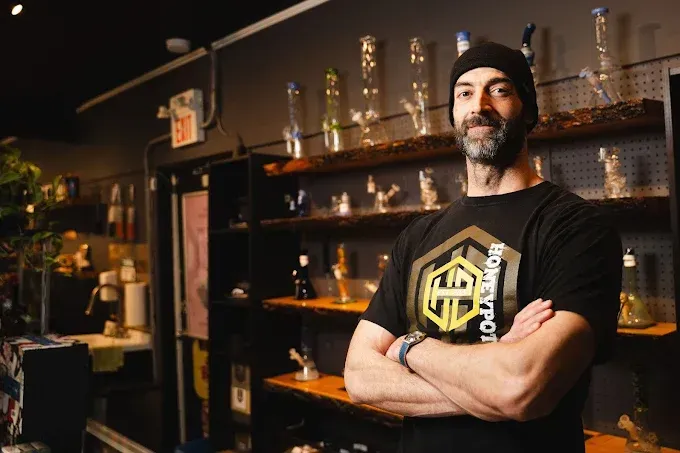 A person with a beard and black cap stands with arms crossed in front of a shop shelf lined with glass smoking accessories in a dispensary in Niagara Falls