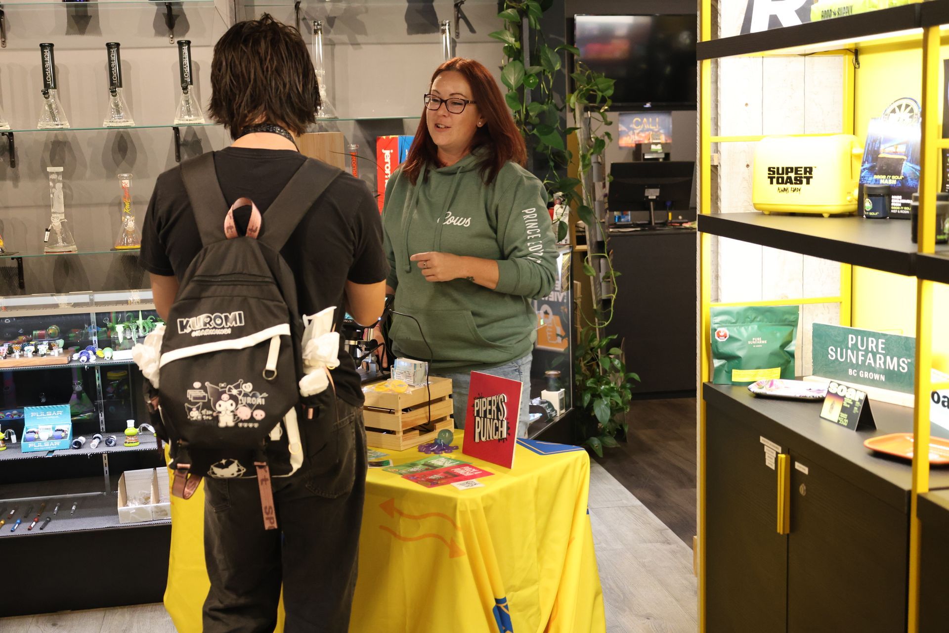 A person wearing a black backpack talks to a staff member in a green hoodie behind a yellow display table in a shop.
