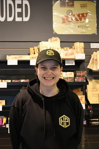 A smiling person wearing a black cap and hoodie with yellow logos, standing in a shop with shelves of smoking products in a niagara falls dispensary